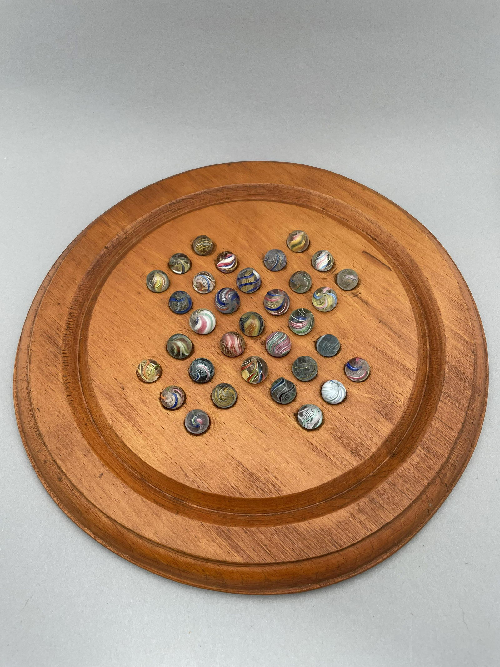 Solitaire Board with Thirty-Three Pee Wee Marbles: The marbles handmade and comprising swirls. Approximate range of diam: 29/64 to 73/128 in. Board: Diam: 10.25 in., D: .75 in.