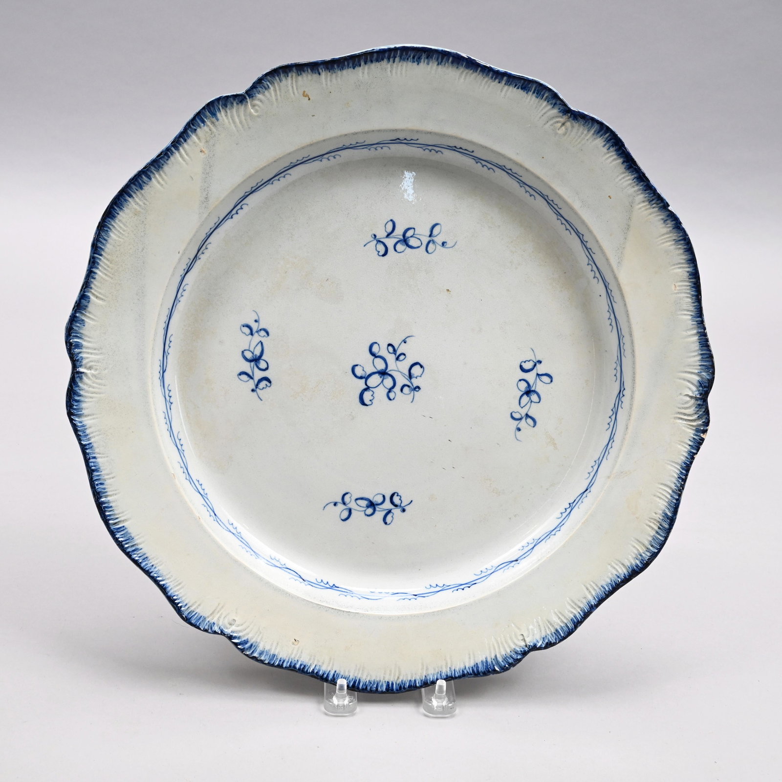 English Pearlware Charger, Circa 1810: Staffordshire or Yorkshire. Painted in blue in the center with a leafy sprig bearing buds, within four smaller sprigs, the well surrounded by a band entwined with vinery, the molded shell edge heighte