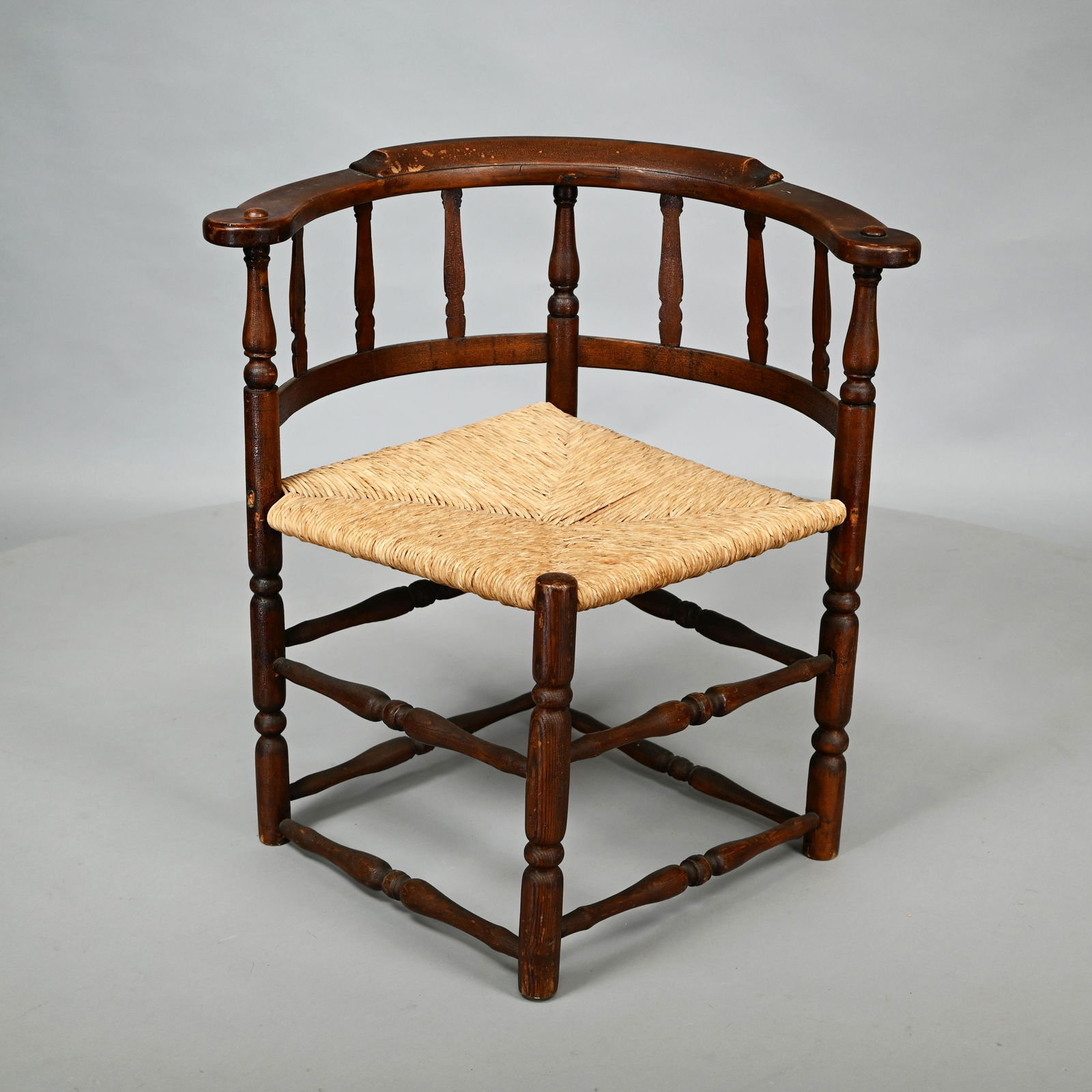 New England Banister-Back Corner Chair: Mid-Eighteenth Century. The curved crest rail above six bannister splats, the rush seat raised on sausage-turned legs joined by double ring-turned vase-form stretchers. Provenance: Found in Hampton, N
