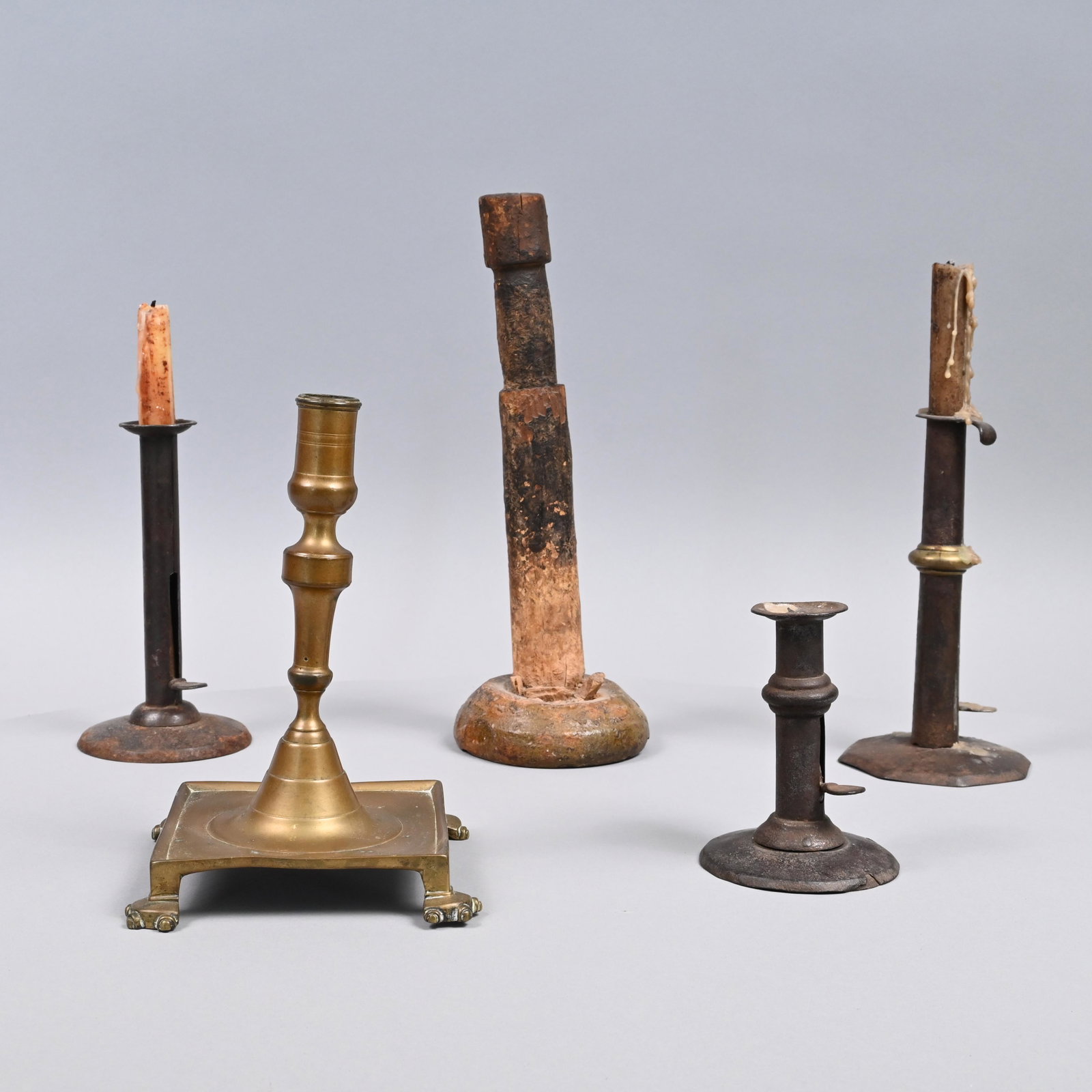 Five Early Brass, Tin and Wood Lighting Devices: Comprising a Spanish brass candlestick, seventeenth century; three tin hogscraper push-up candlesticks, one with brass fitting; and a turned wood candlestick mounted in a glazed green and orange redwa