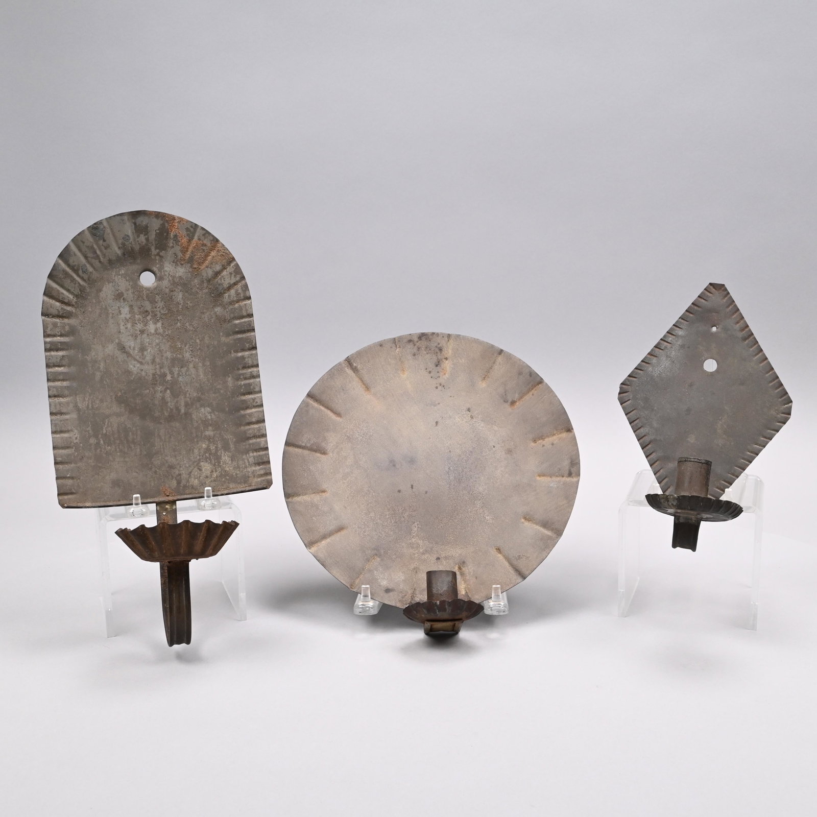 Three American Tin Single-Light Sconces: Nineteenth Century. The backplates crimped and comprising one diamond-shaped, one arched and one circular backplate, each fitted with a cylindrical nozzle above a crimped drip-pan. Diamond sconce: H: 