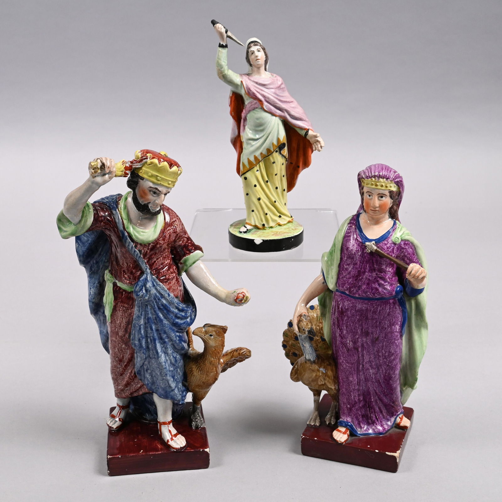 Three Staffordsdshire Pearlware Figures, 1810-20: Comprising a pair, well-colored in enamels and modelled as 'Jupiter' attended by his eagle, and 'Juno' by her peacock, each on a square brown base; the third a woman holding a dagger, on a circular ba