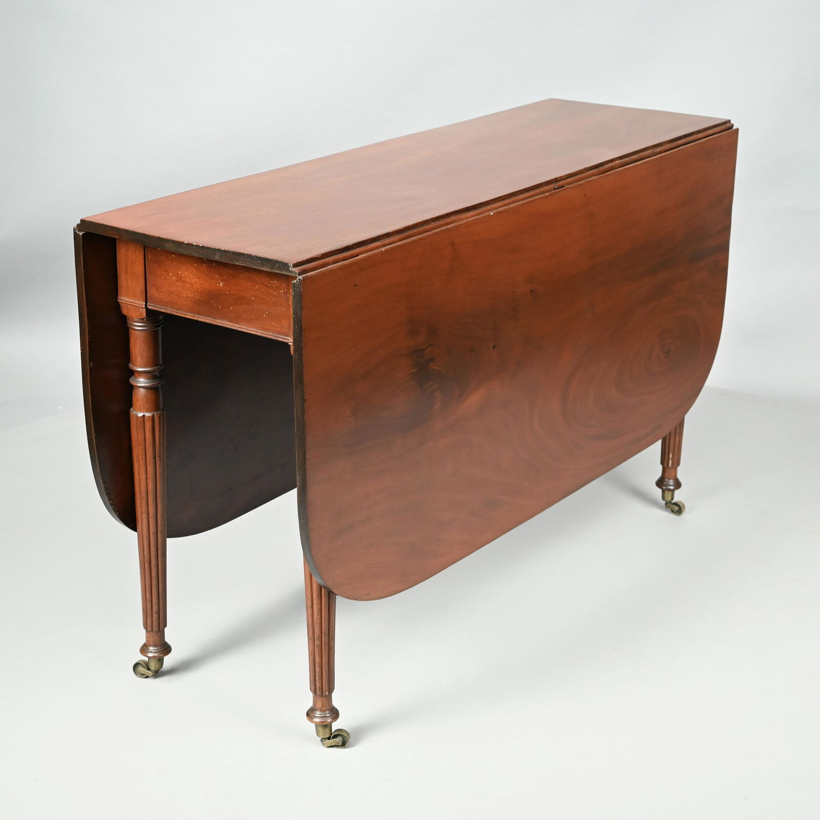 Sheraton Mahogany Drop-Leaf Dining Table: Early Nineteenth Century. The oblong top above rounded leaves and a plain frieze with molded edge, raised on ring-turned and slightly tapered reeded legs ending in cuffs, on brass casters. H: 29.25 in