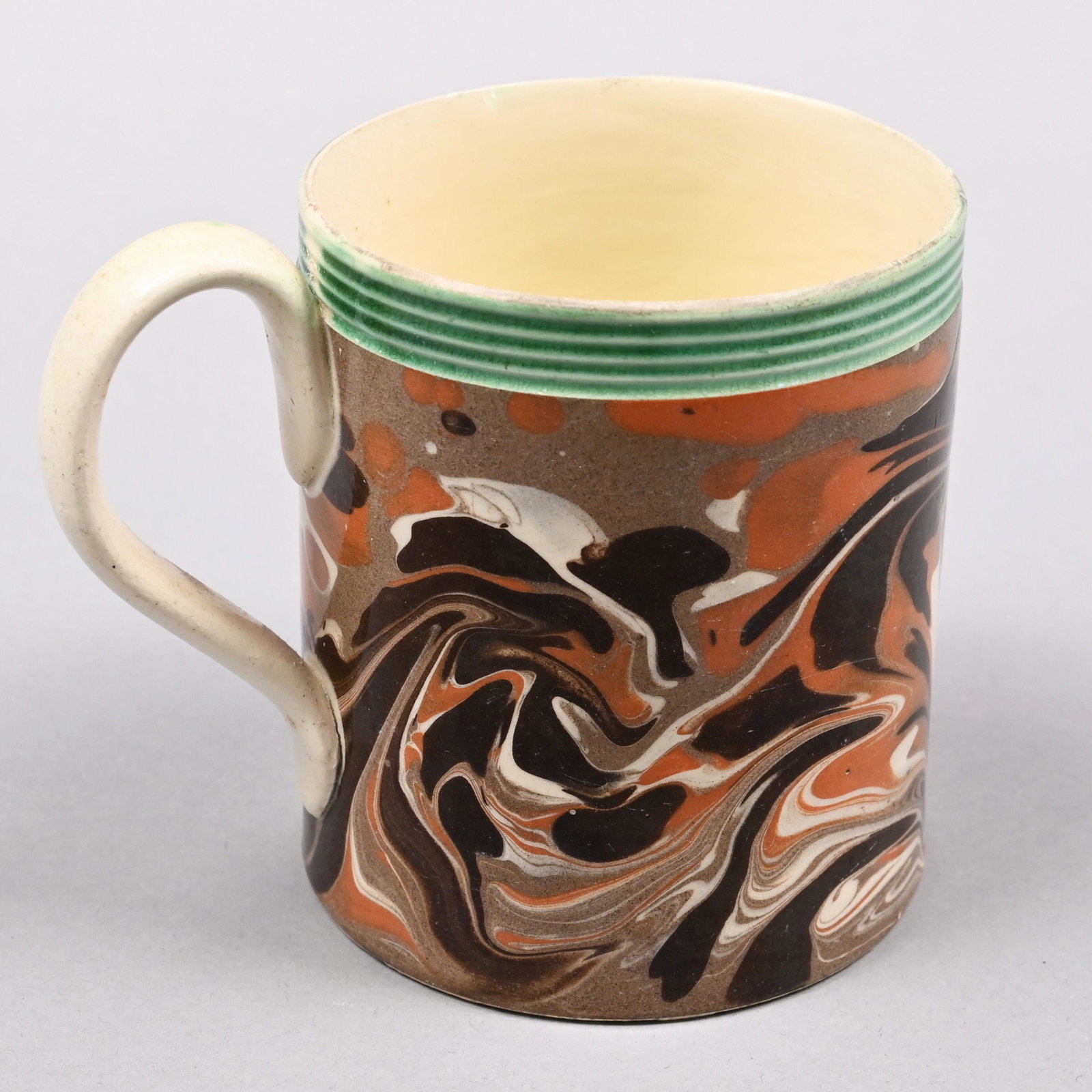 British Creamware Mochaware Mug, 1790-1800: Slip-marbled in black, brown, pumpkin and white on a taupe field, beneath a rouletted green-glazed reeded border. H: 3.375 in., W: 2.875 in., D: 4.125 in.