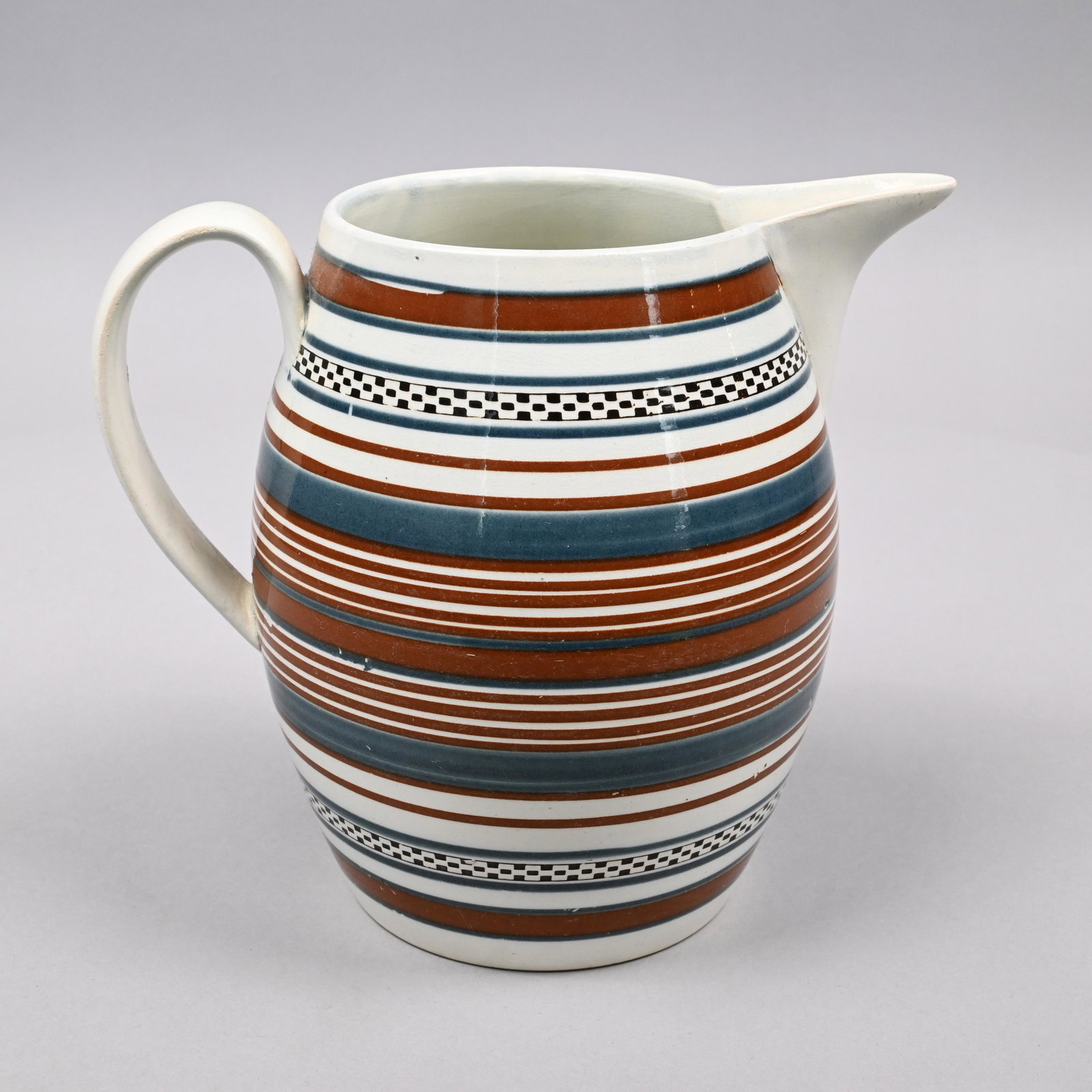 British Pearlware Mochaware Jug, Circa 1820: Surrounded by two rouletted black slip-filled checkered bands, within turned graduated rust and slate blue bands. H: 8.25 in., W: 6 in., D: 9.25 in.