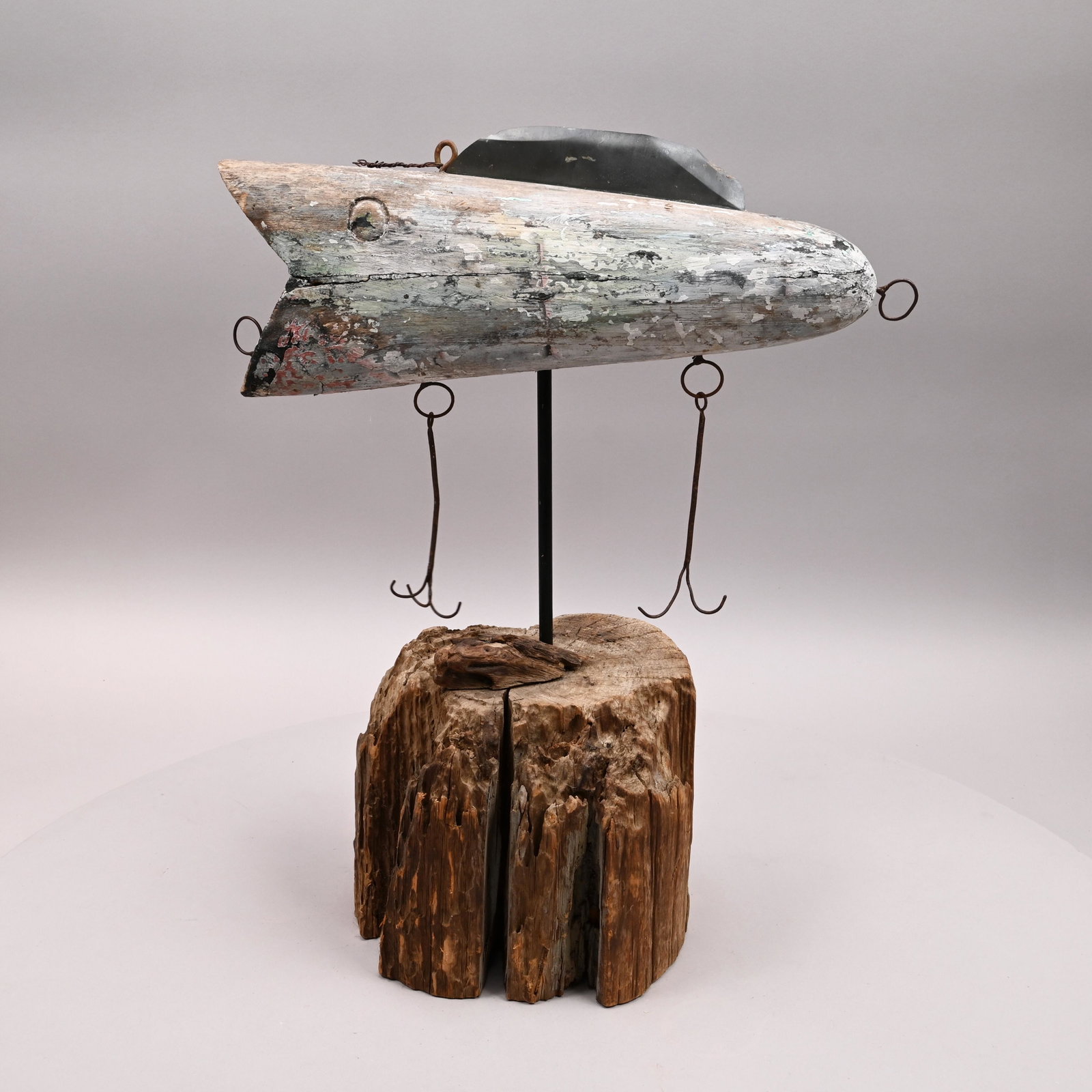 Folk Art Carved & Painted Figure of a Fishing Lure: Twentieth Century. In traces of blue-green and white paint, incised circular eye, wearing a tin fin and suspending two large hooks, mounted on a driftwood stump. H: 22.25 in., W: 18.25 in., D: 10 in.