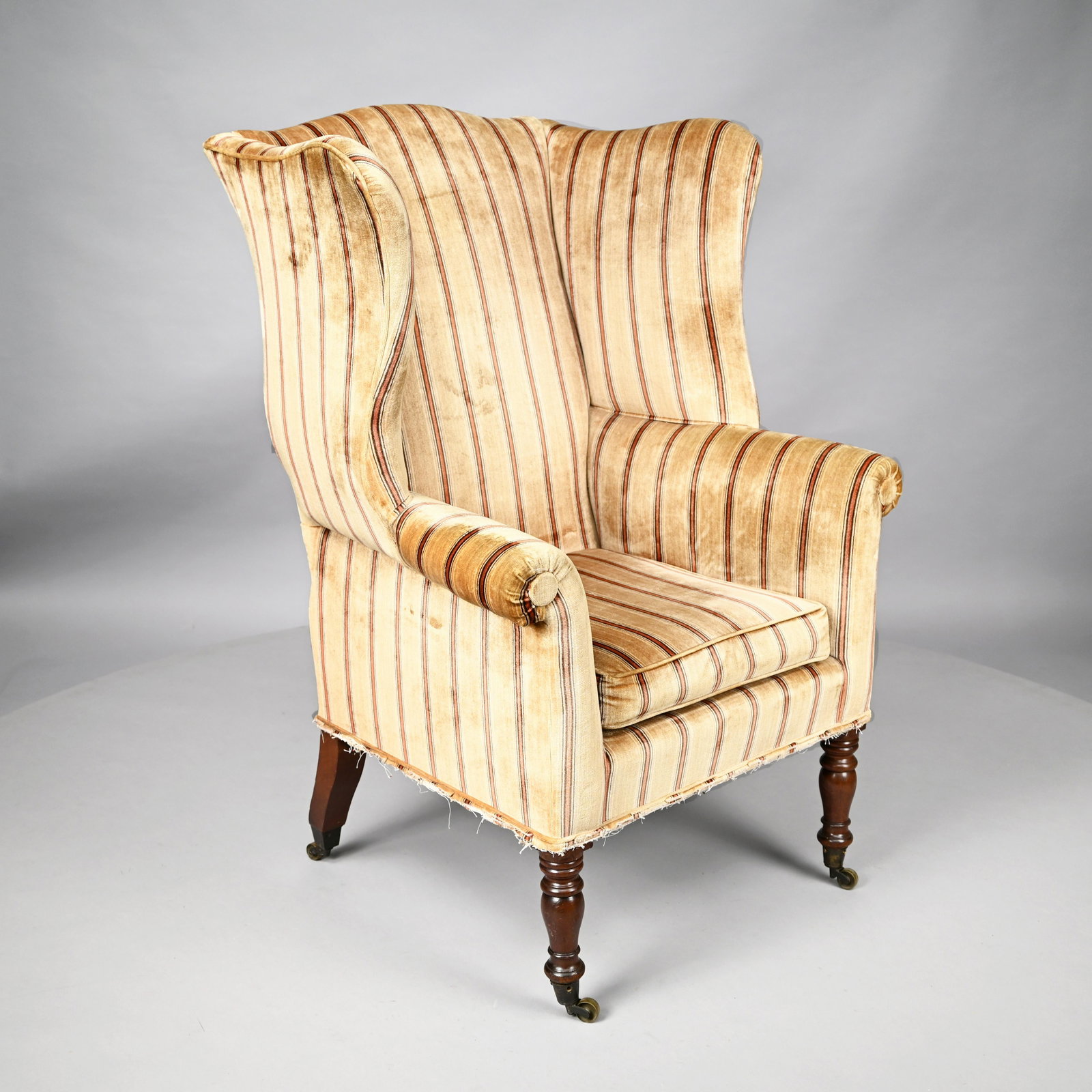 Sheraton Mahogany Wingback Armchair: Early Nineteenth Century. Of typical form with out-scrolled arms, the loose seat cushion above the apron and raised on frontal ring-turned swelled legs, upholstered in striped brushed velvet, on caste