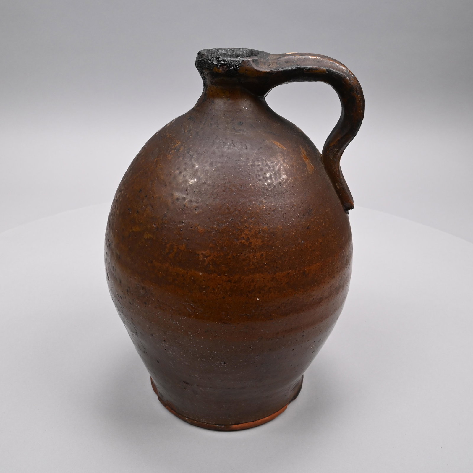 New England Glazed Redware Jug: First-Half Nineteenth Century. The turned jug with waisted neck and footed base, applied ridged handle, covered in dark brown manganese glaze. H: 13 in., W: 8 in., D: 8 in.