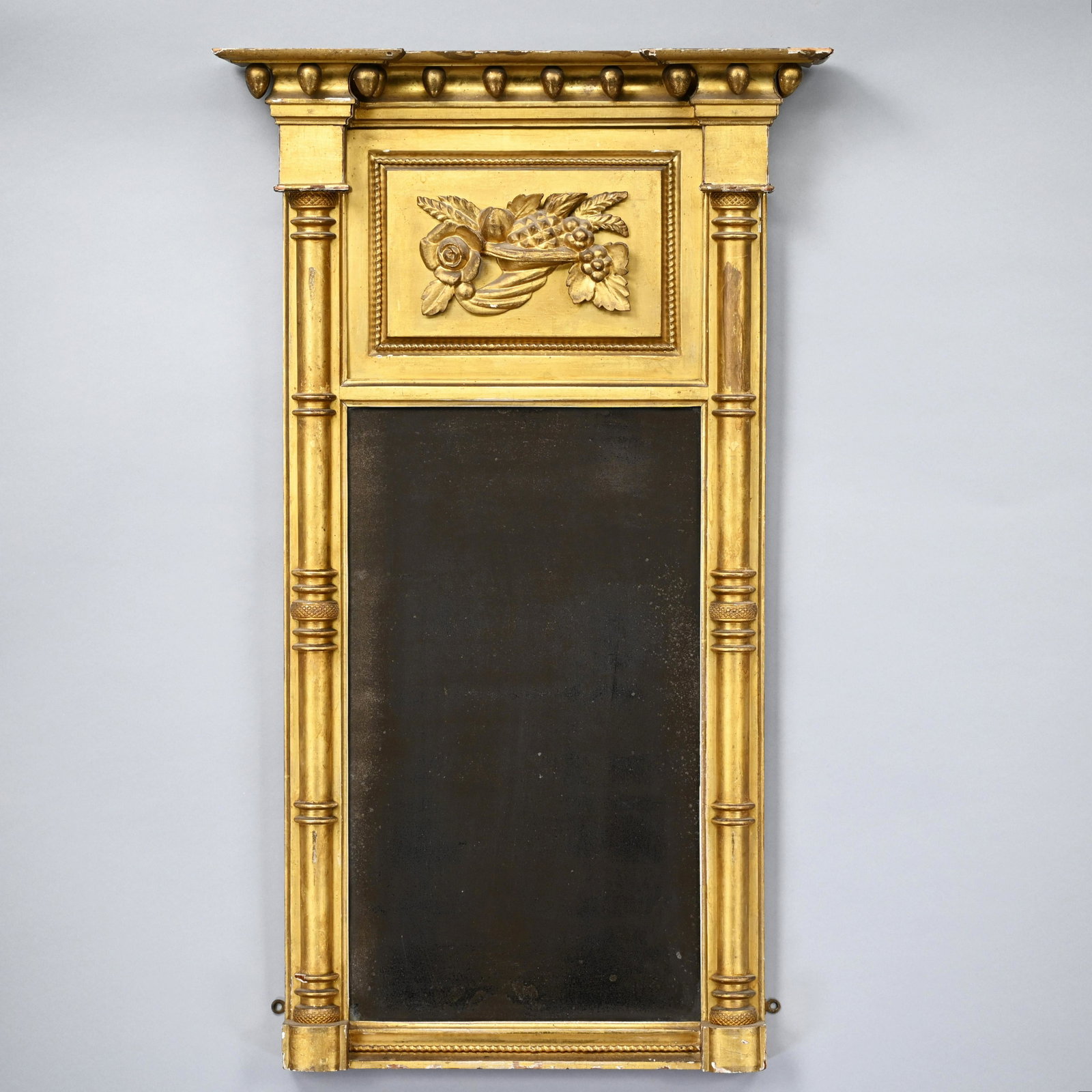 Classical Giltwood Tabernacle Mirror, Circa 1830: The rectangular cornice with outset corners hung with oval pendants, above a raised panel applied with a carved cornucopia filled with fruit and flowers, over a rectangular mirror plate, within engage