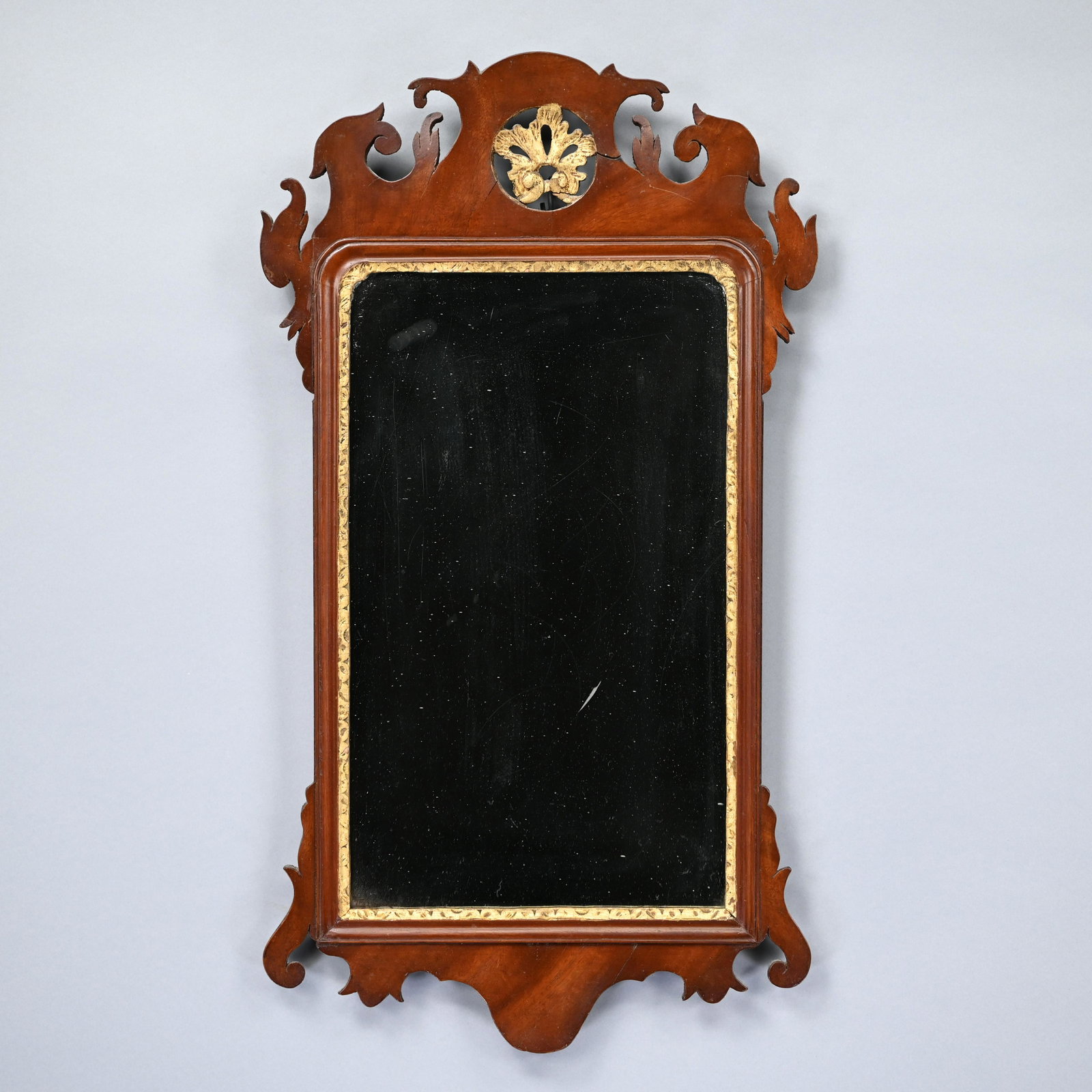 Chippendale Mahogany and Parcel-Gilt Looking Glass: The rectangular mirror plate within a gilt surround, the scroll crest centered by a roundel applied with a rococo pierced gilt shell, above a matching scroll apron. H: 30.875 in., W: 17 in., D: 1.75 i