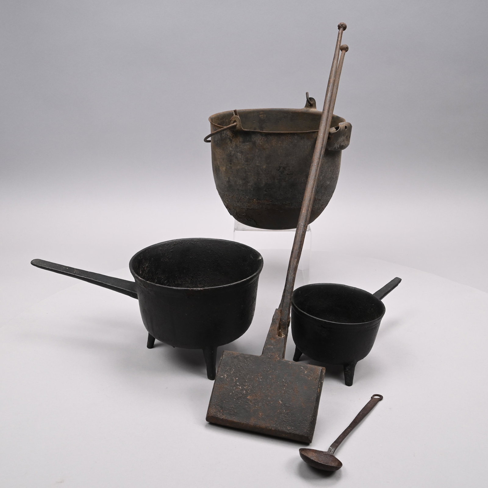 Four American Cast & Wrought-Iron Cooking Utensils: Late Nineteenth-Twentieth Century. Comprising two graduated posnet pots, each on three feet; a waffle iron cast in a grid pattern; and a ladle. Together with a steel kettle with swing handle, Bolh Ste