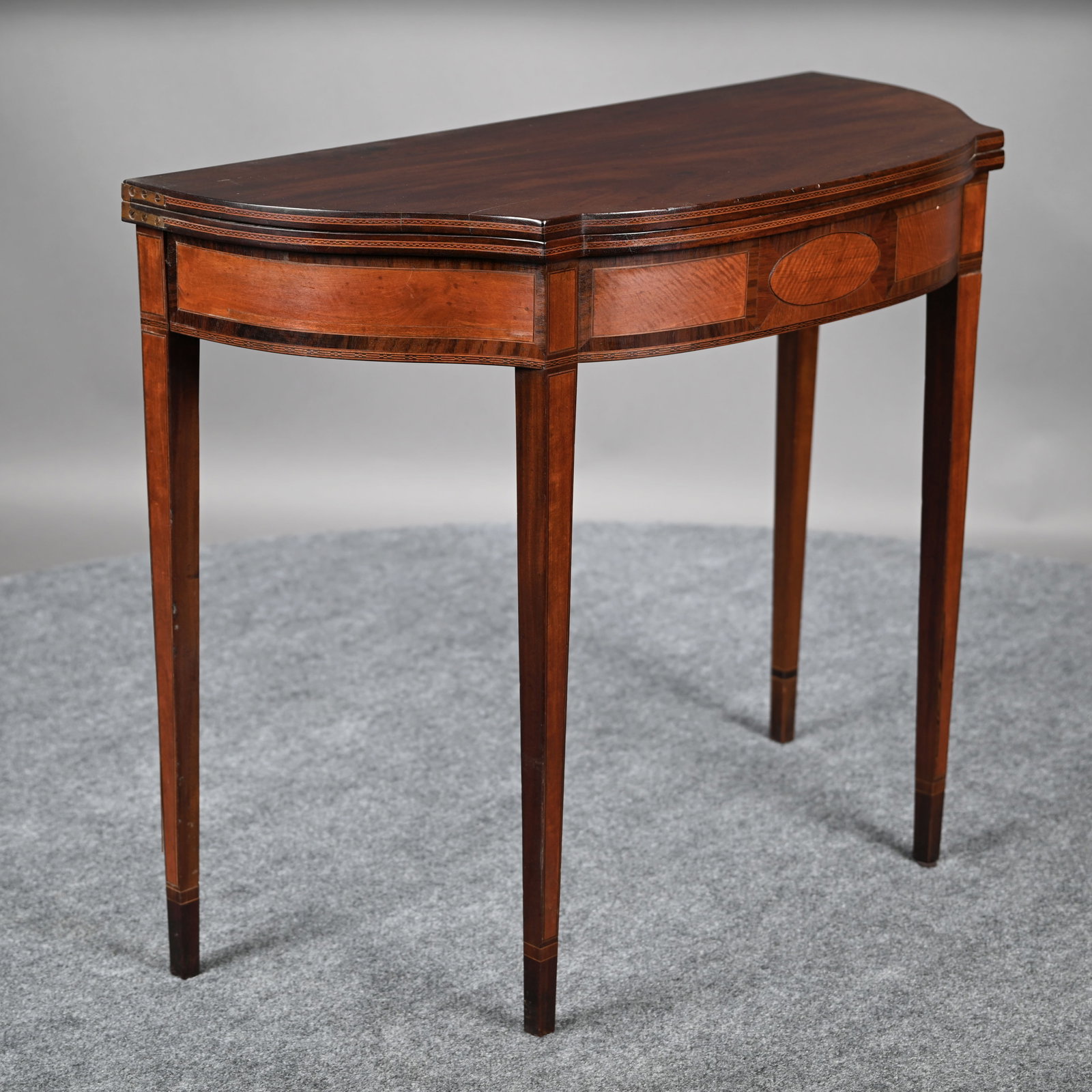Federal Inlaid Mahogany Card Table: Federal Inlaid Mahogany Card Table. The D-shaped top above the frieze inlaid with an oval patera, opening to a circular plain playing surface with notched corners, raised on square tapered line-inlaid