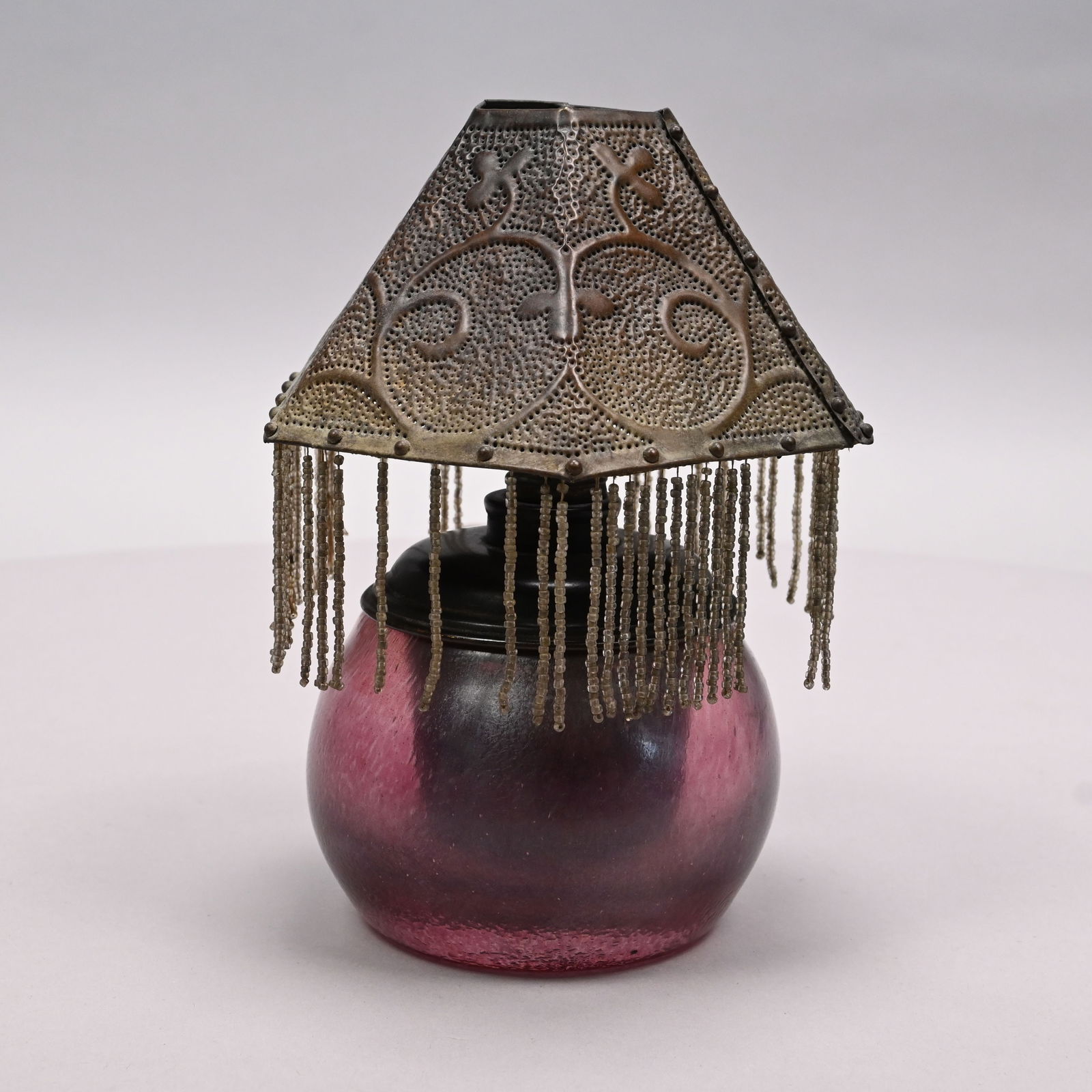 Art Glass Oil Lamp with Embossed Brass Shade: Art Glass Oil Lamp with Embossed Brass Shade, 1900-20. The oil lamp in dark rose pink, engraved "Loetz/ Austria," the shade embossed with scrolling vinery and suspending beaded chains. Base: H: 4.25 i