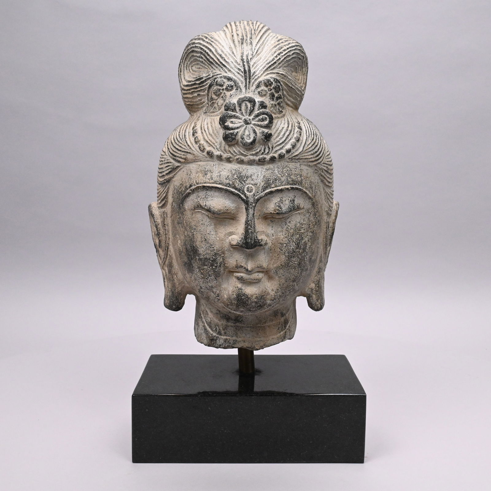 Carved Stone Head of Buddha: Carved Stone Head of Buddha. Serene visage, wearing a topknot (ushnisha) representing wisdom and enlightenment, and adorned with a lotus flower and row of tight curls.Head: H: 13.5 in., W: 8.125 in.,