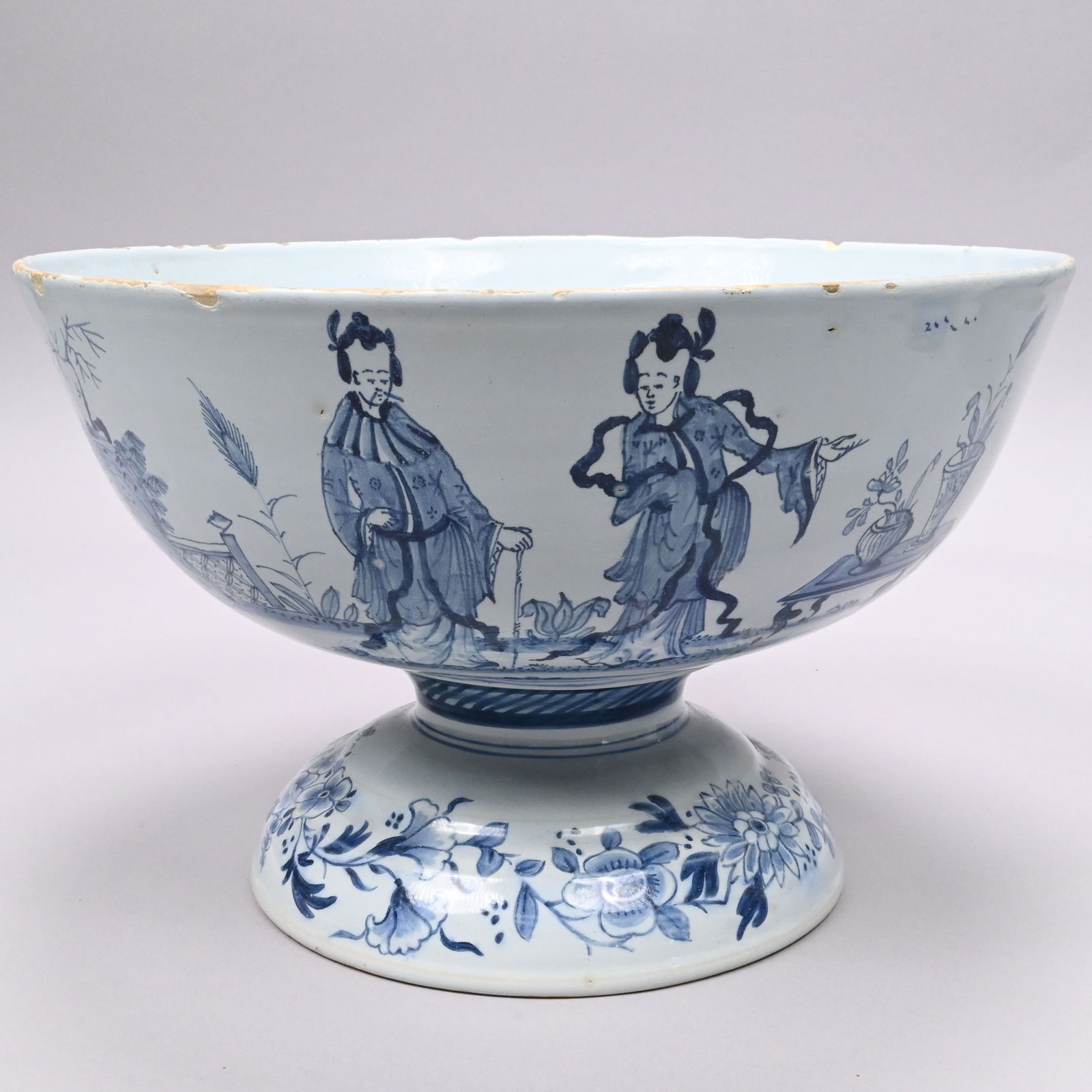 English Delft 'Ribbon' Chinoiserie Pedestal Bowl: English Delft Blue and White 'Ribbon' Chinoiserie Pedestal Punch Bowl, Circa 1750, Probably Bristol. Painted around the sides with two vases of flowers on a low table beside a basket and scroll,