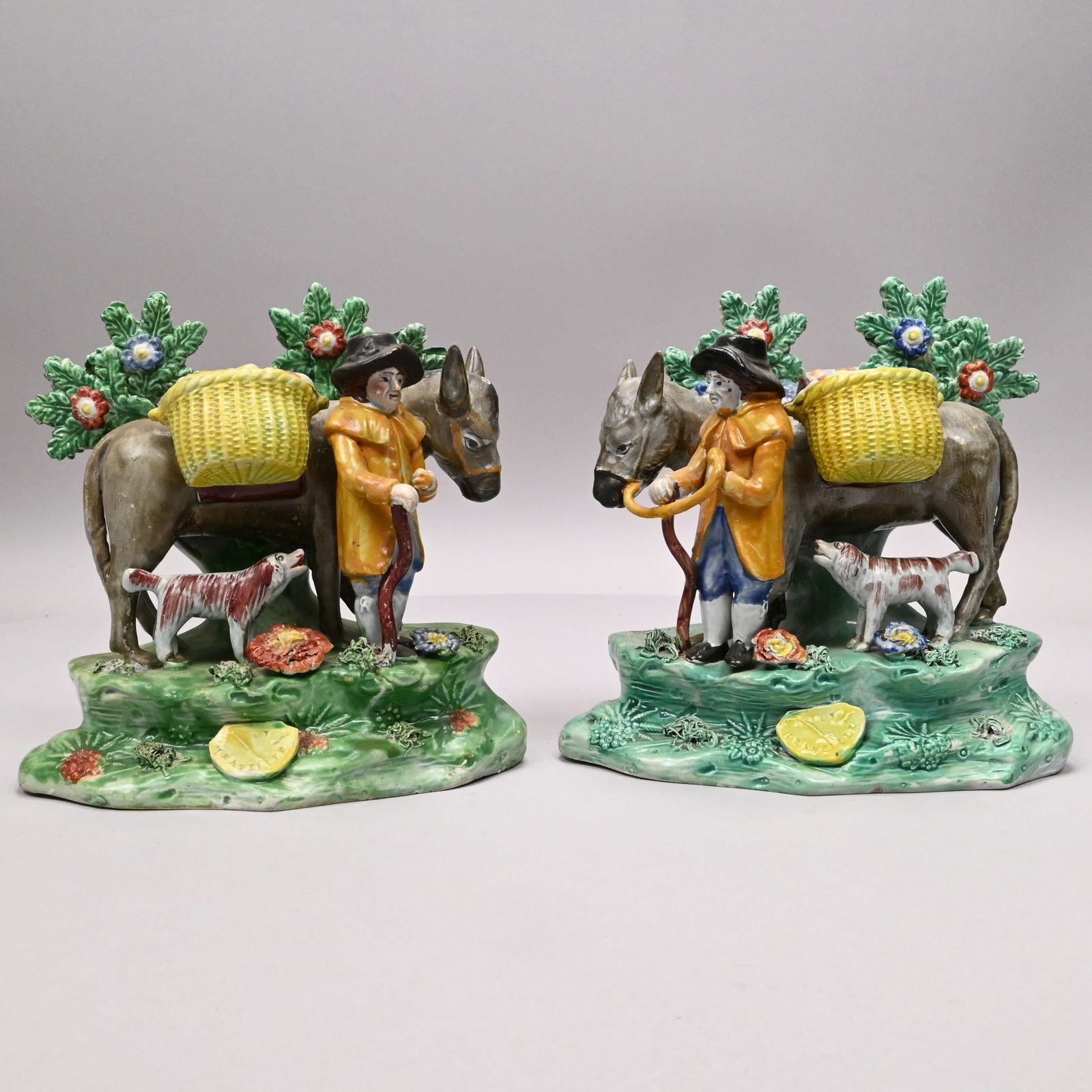 Two Staffordshire 'Jolly Traveller' Figural Groups: Two Staffordshire Pearlware Figural Bocage Groups of the 'Jolly Traveller,' Circa 1820. Each man leading a mule laden with two baskets filled with produce, accompanied by his dog behind him, raised