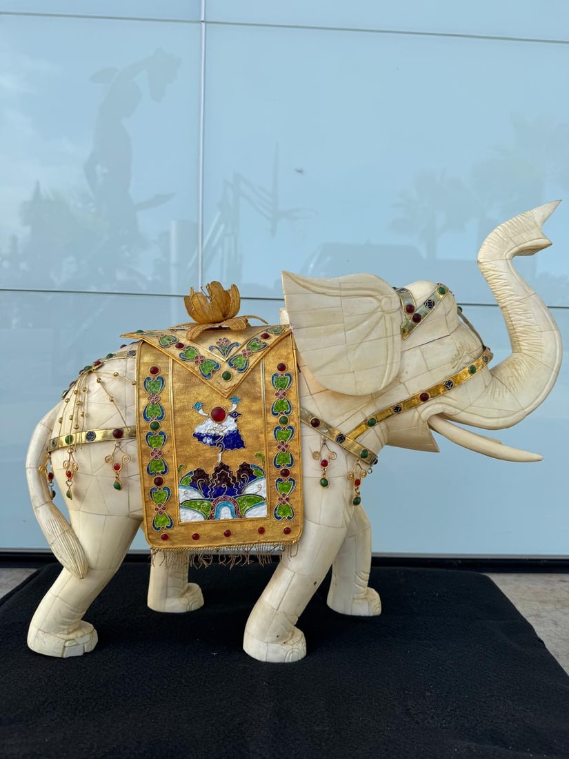 Hand-Carved Jeweled Elephant Sculpture with Enamel and Gold Detailing: Size:26Lx20hx9wA beautifully handcrafted elephant sculpture carved from a light, ivory-toned material, adorned with an ornate golden saddle blanket richly detailed with colorful enamel work and