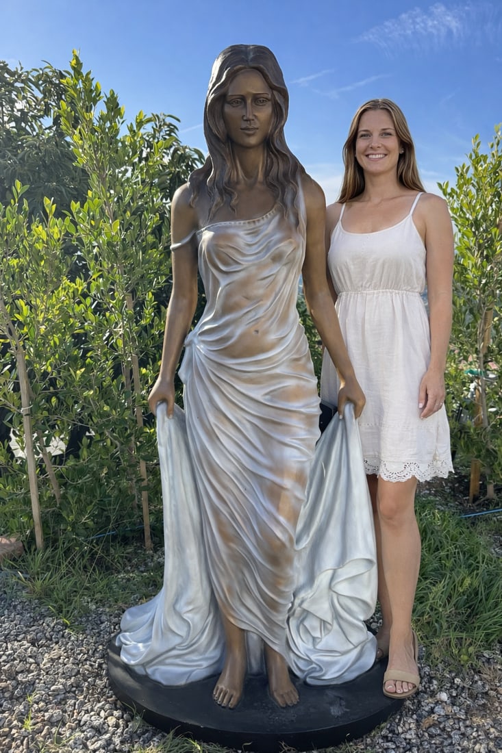 Life Size Bronze Lady Sculpture: Size: 70” x 37”This elegant sculpture depicts a serene female figure in a classical Greco-Roman–inspired style. She is shown standing barefoot on a round base, gently holding the