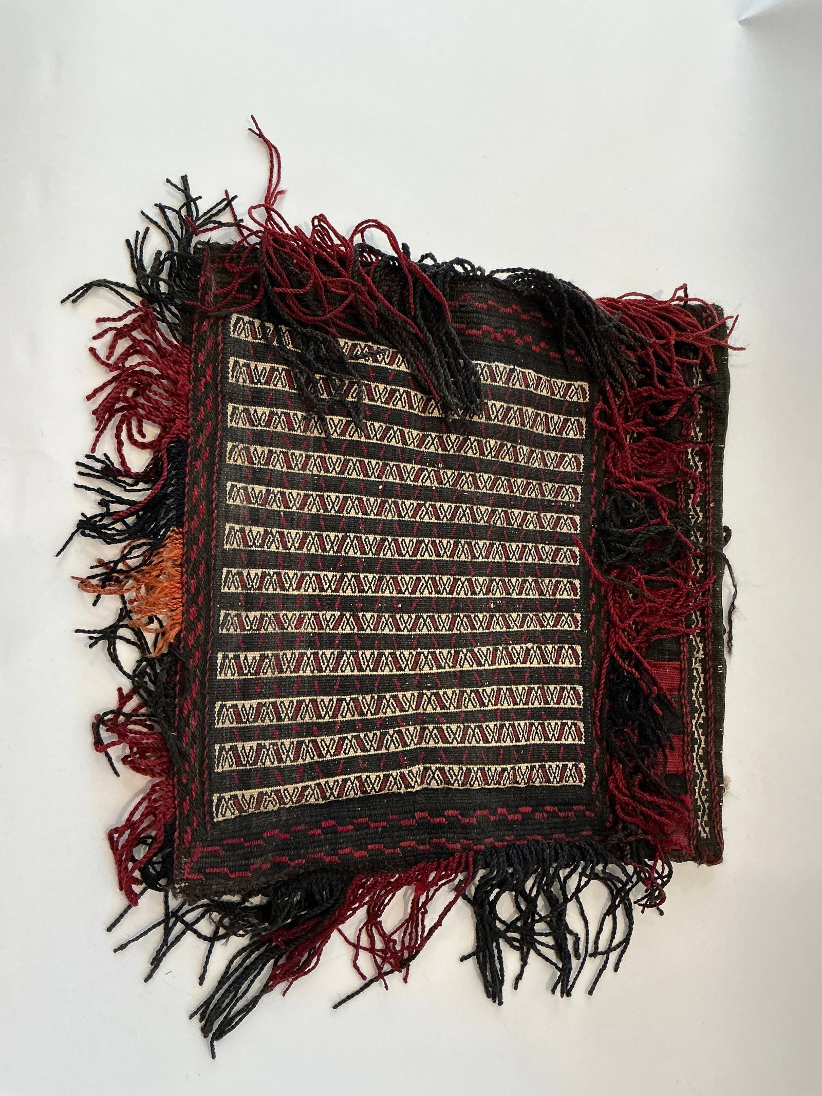 Vintage Handwoven Wool Camel Bag from Pakistan: A vintage handwoven wool camel bag from Pakistan, showcasing traditional geometric patterns in rich red and black hues, accented with cream. This piece is crafted from 100% wool and features fringed e