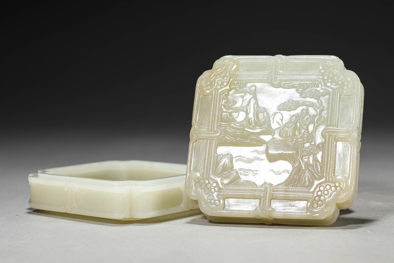A JADE 'CARVED SCHOLARS UNDER A PINE TREE' 'INDENTED CORNER' COVERED BOX: A JADE 'CARVED SCHOLARS UNDER A PINE TREE' 'INDENTED CORNER' COVERED BOX Dimensions: Width: 9.0 cm (3.54 in) x Height: 3.6 cm (1.42 in). All measurements are approximate.