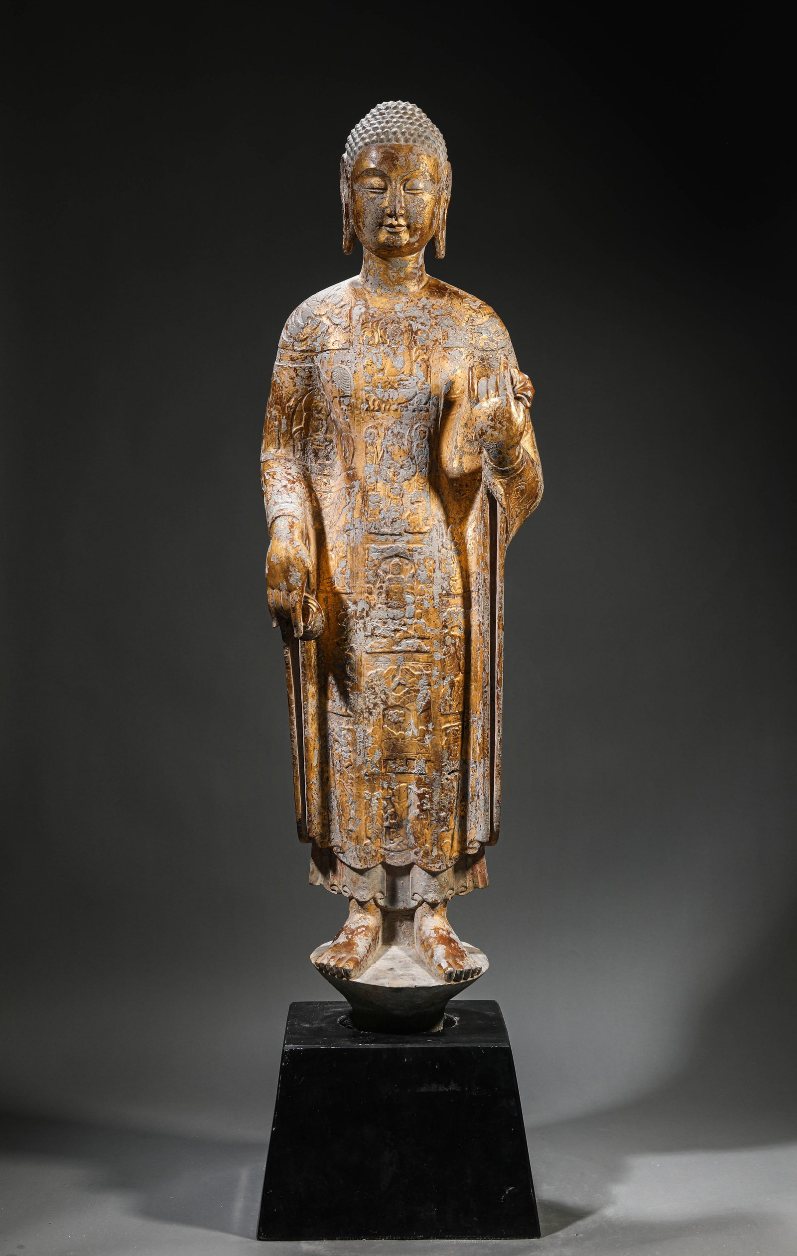 A STONE AND GILT-LACQUERED STANDING FIGURE OF SHAKYAMUNI: A STONE AND GILT-LACQUERED STANDING FIGURE OF SHAKYAMUNI Dimensions: Length: 30.0 cm (11.81 in) x Width: 28.0 cm (11.02 in) x Height: 115.5 cm (45.47 in). All measurements are approximate.