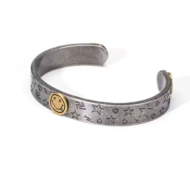 NIRVANA SMILEY FACE CUFF BRACELET: NIRVANA SMILEY FACE CUFF BRACELET Description: Cuff bracelet in a silver-toned metal with an open-ended form and engraved allover motifs including stars, hearts, lightning bolts, and a swastika