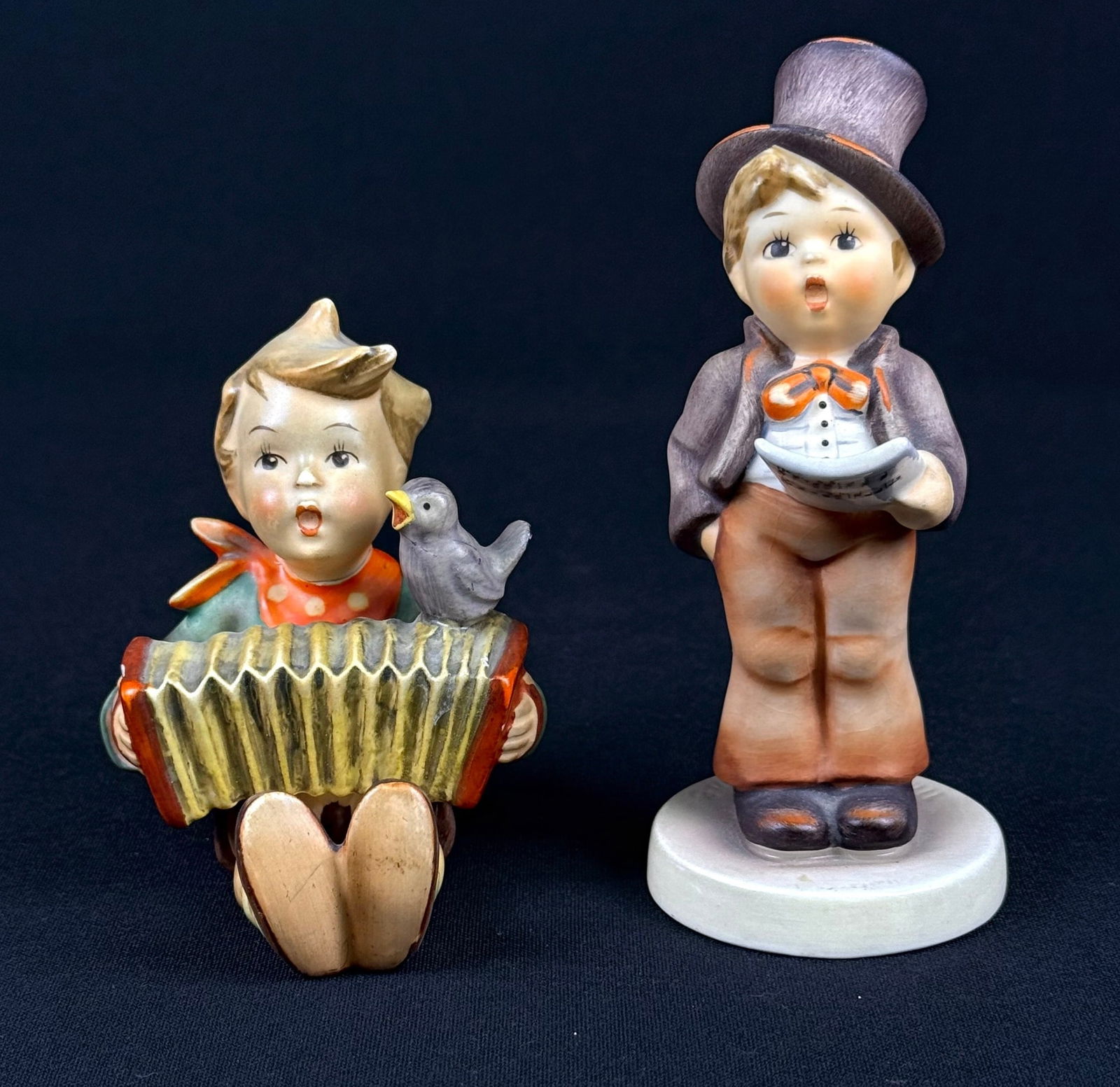 Pair of Goebel M.I. Hummel Figurines: Heini, Bandoneon Player (Model 110/0) & Let's Sing (Model 137): A pair of glazed ceramic or porcelain figurines by Goebel M.I. Hummel. The first figure, identified as 'Heini, Bandoneon Player' (Model 110/0), depicts a stylized child figure seated, playing a yellow
