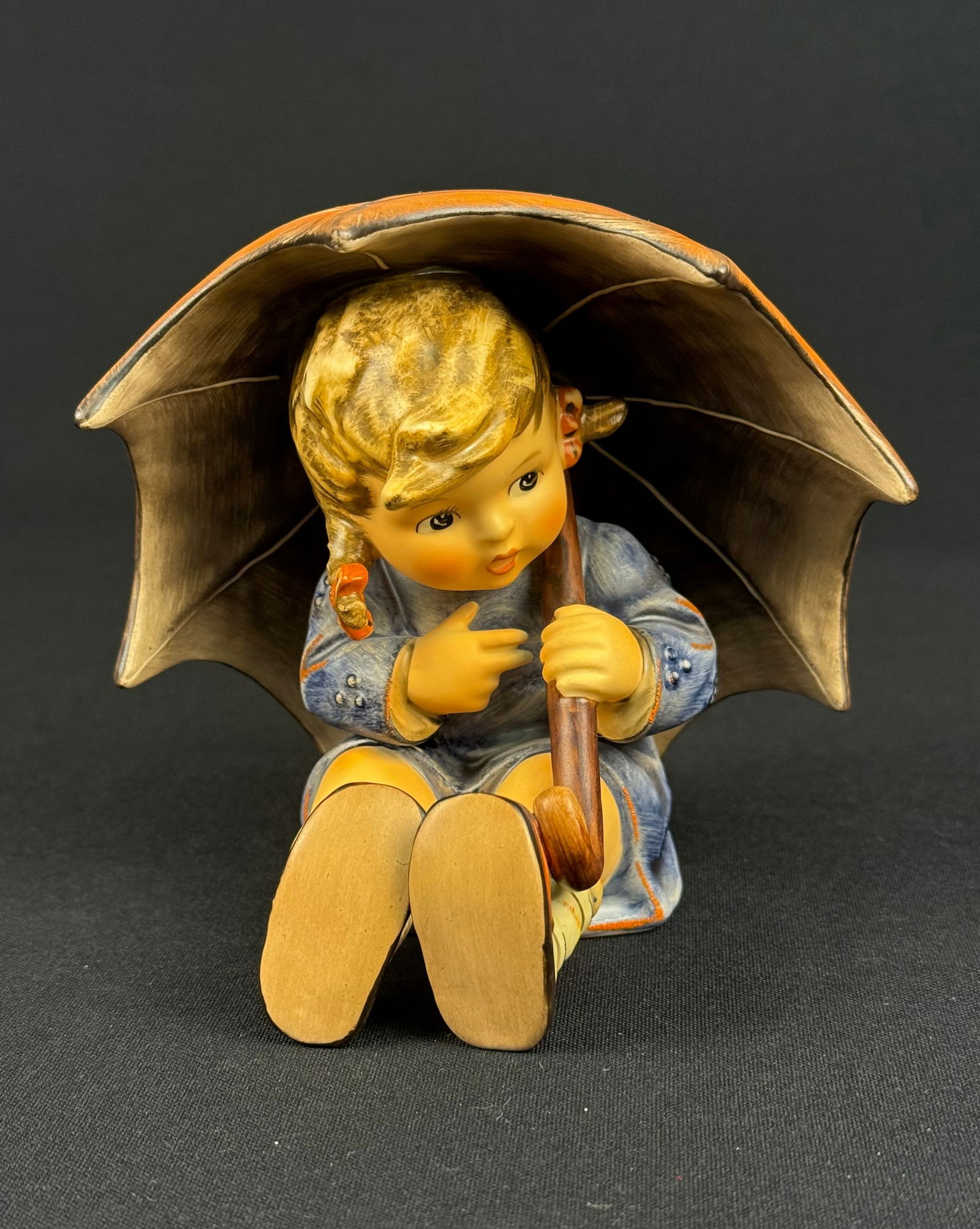Goebel Hummel 'Umbrella Girl' Figurine, Model 15270 B, 1957: A Goebel Hummel "Umbrella Girl" figurine, model 15270 B. This glazed ceramic piece depicts a young girl seated beneath a two-toned orange and brown umbrella. The figurine features an embossed "M. I. H
