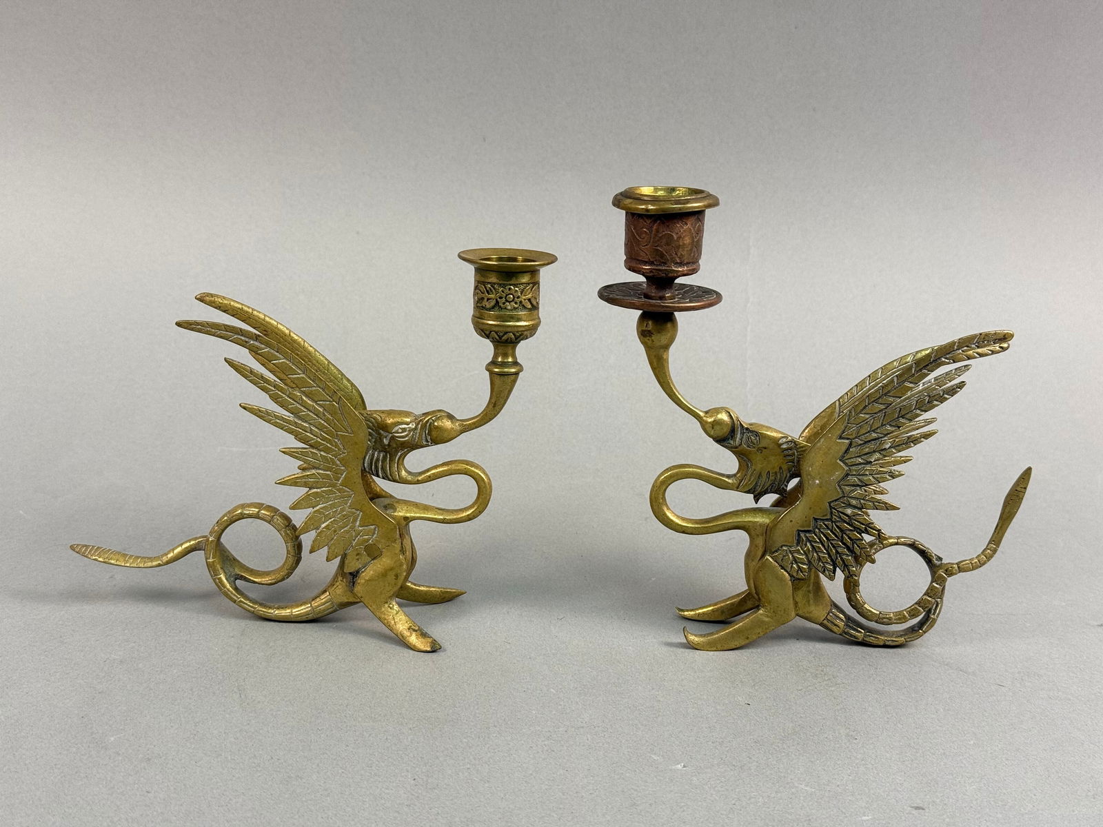 A Pair of Late 19th / Early 20th Century Revivalist Brass Dragon / Griffin Candlesticks: A pair of revivalist brass candlesticks, each crafted as a winged, serpent-like creature or griffin supporting a decorative candle cup. The pieces feature ornate detailing on the creature's wings, sca