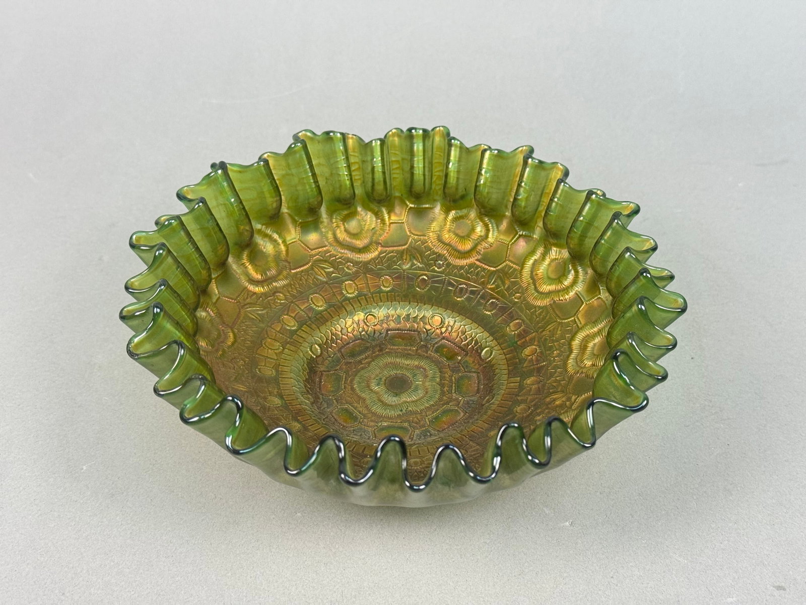 Fenton Green Carnival Glass Crimped Ribbon Bowl: A Fenton green Carnival Glass bowl. This round, footed bowl is crafted from pressed glass and features a deeply ruffled or crimped rim. The interior design is composed of concentric bands of geometric