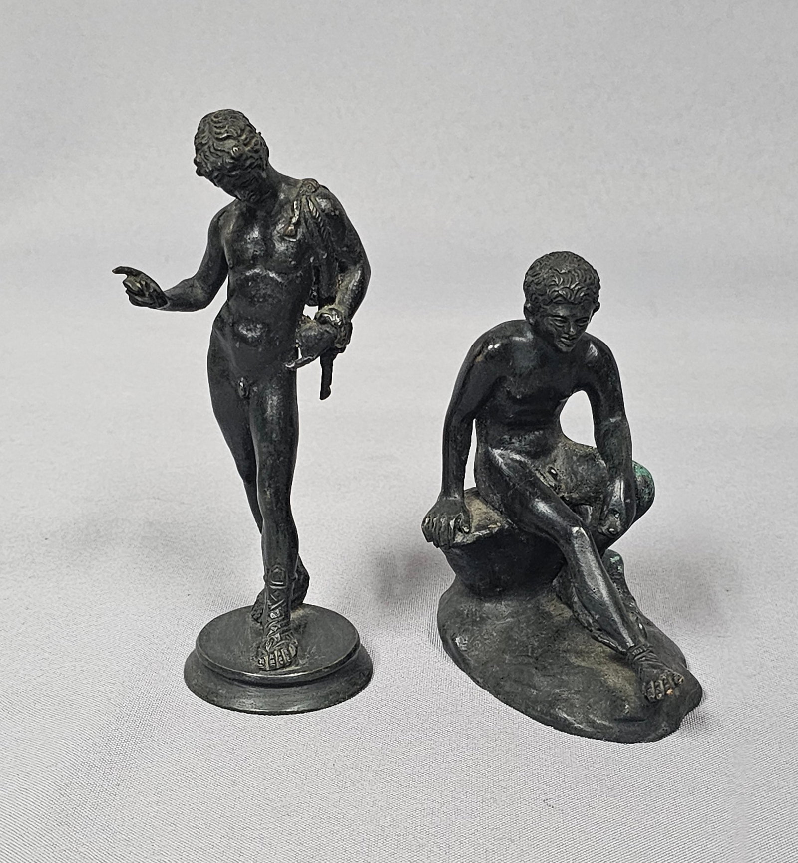 A Pair of Patinated Bronze Figures, including a Seated Hermes: This lot comprises two patinated bronze figurines depicting nude male figures in a classical or neoclassical style. The first figure, identified as 'Seated Hermes' (after the Antique, attributed to Ly