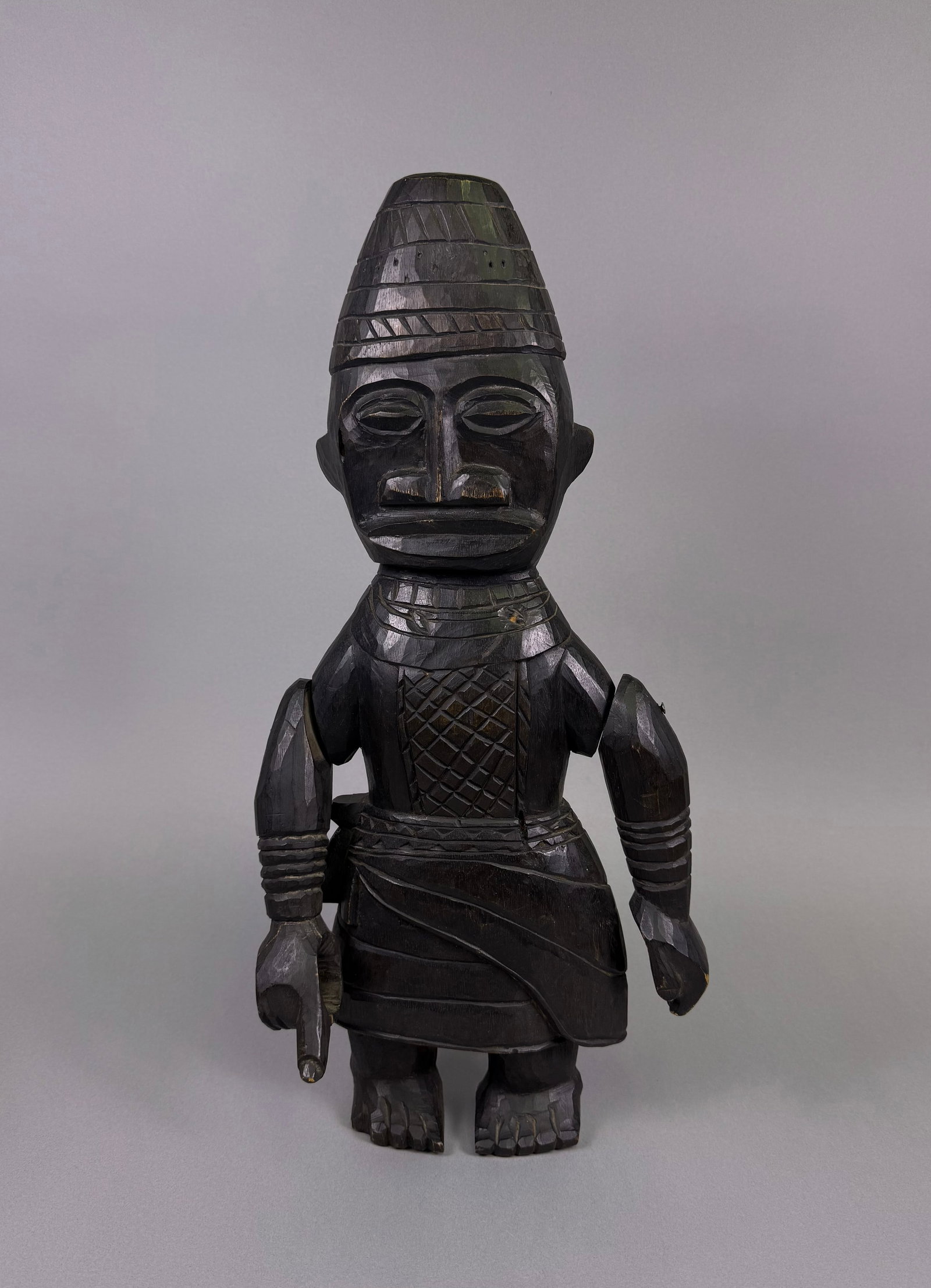 A Yoruba Ibeji Twin Figure, Male, Carved Wood: A Yoruba Ibeji Twin Figure, Male, carved from dark wood, presenting as a standing, stylized anthropomorphic figure. It wears a conical, ribbed headdress and a skirt-like lower garment. The arms are ar
