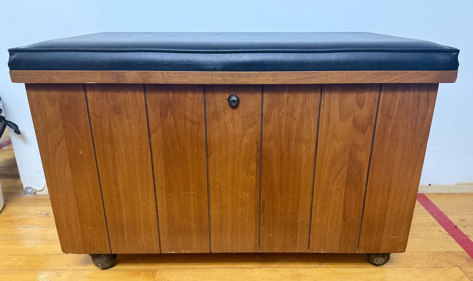 Lane Mid-Century Modern Record Storage Bench: A Mid-Century Modern record storage bench by Lane, constructed from finished wood with a vertically slatted front and resting on small brass-toned casters. The hinged lid is topped with a black padded