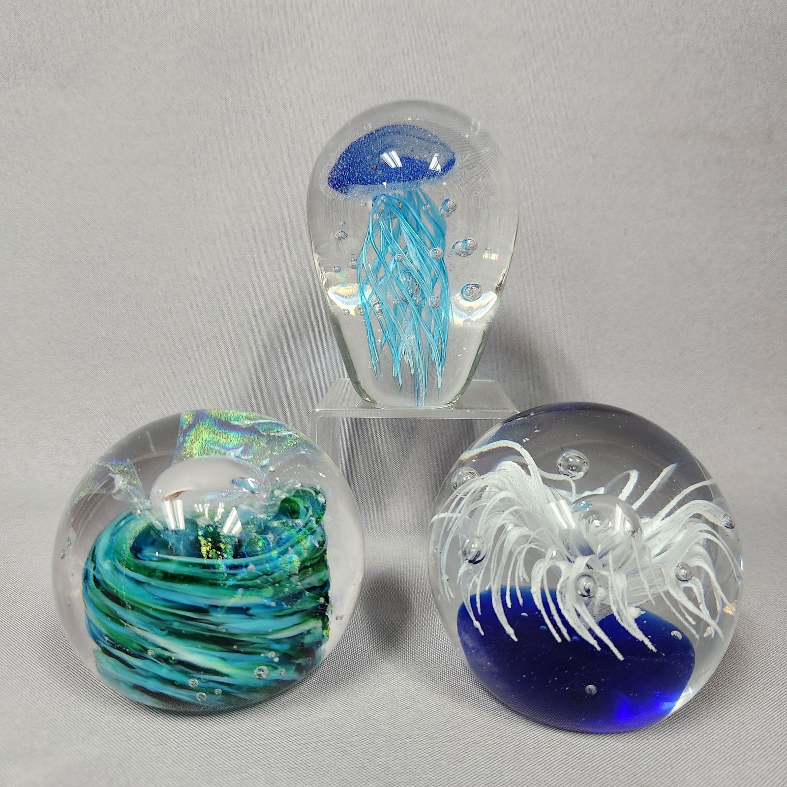 Three Contemporary Art Glass Paperweights, including a 'Jellyfish' example and a signed 'Dai Watts': This lot comprises three contemporary art glass paperweights. The first is a spherical form, crafted from clear glass encasing an iridescent swirl of green, blue, and black glass with a central clear