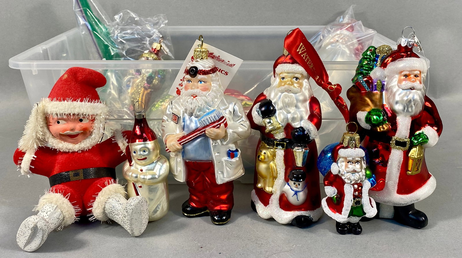 A Collection of Christmas Ornaments: A collection of Christmas ornaments, predominantly featuring figures of Santa Claus, alongside a few snowmen and other thematic variations. Among the collection is a blown glass 'Doctor Santa' ornamen