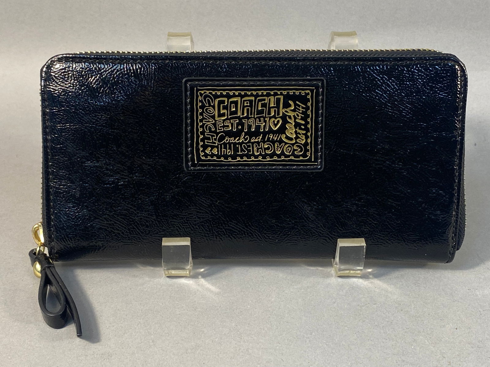 Coach Poppy Black Patent Leather Zip Around Wallet: A Coach Poppy Black Patent Leather Zip Around Wallet. Constructed from a black, shiny, textured material with gold-tone metal hardware. The front features a prominent rectangular emblem embossed with