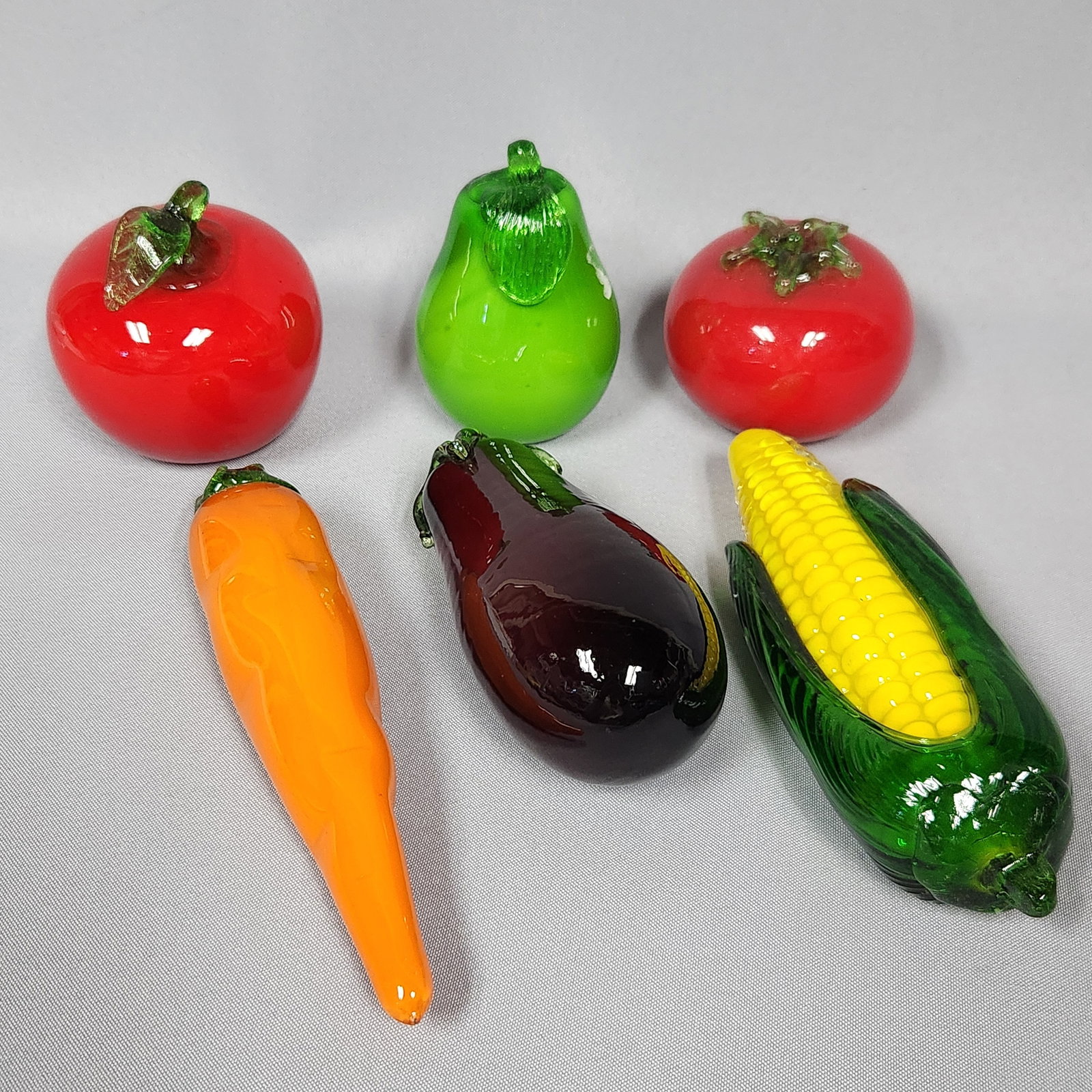 A Set of Six Murano Style Art Glass Fruits and Vegetables: A set of six Murano Style art glass fruits and vegetables, comprising two red tomatoes, one green pear, one orange pepper, one dark purple eggplant, and one yellow and green ear of corn. Each piece is
