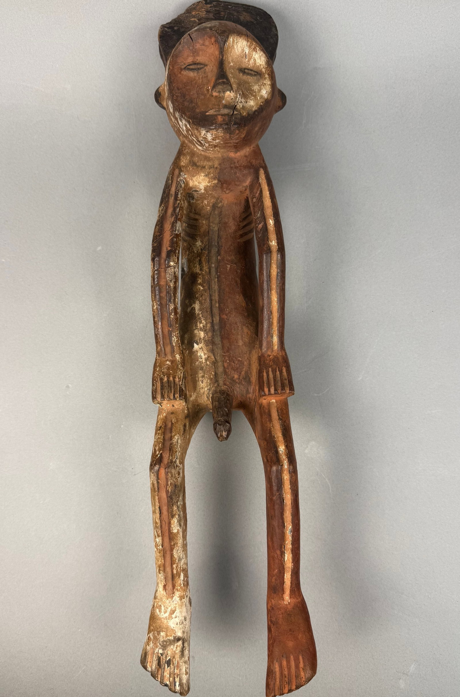 An African Carved Wooden Male Ancestor Figure: A carved wooden anthropomorphic figure, depicting a stylized male form with distinct genitalia, an elongated body with indicated ribs, bent limbs terminating in stylized hands and feet, and a rounded 