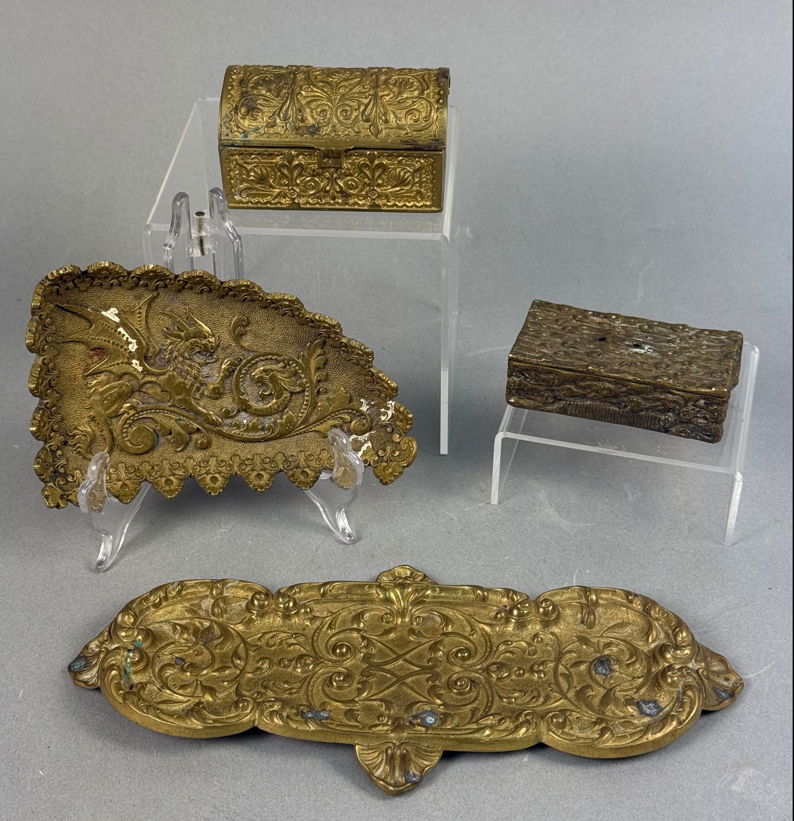 A Group of Vintage Brass Desk Accessories, including Inkwells and Trays: A group of four vintage brass desk accessories. The collection includes a small, rectangular box with a domed, hinged lid, elaborately adorned with rococo-style foliate scrollwork in relief, featuring