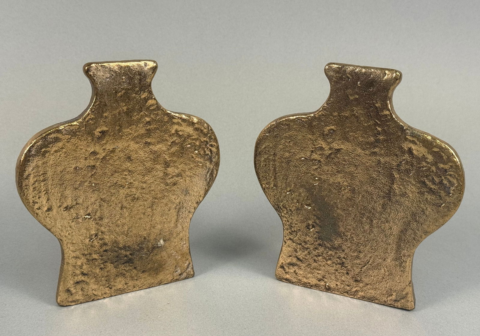 A Pair of Gilt and Patinated Cast Metal Vase-Form Bookends: A pair of cast metal bookends, each presenting a stylized, flattened silhouette reminiscent of an amphora or classical vase. The front surfaces display a textured, uneven gold-toned finish, while the