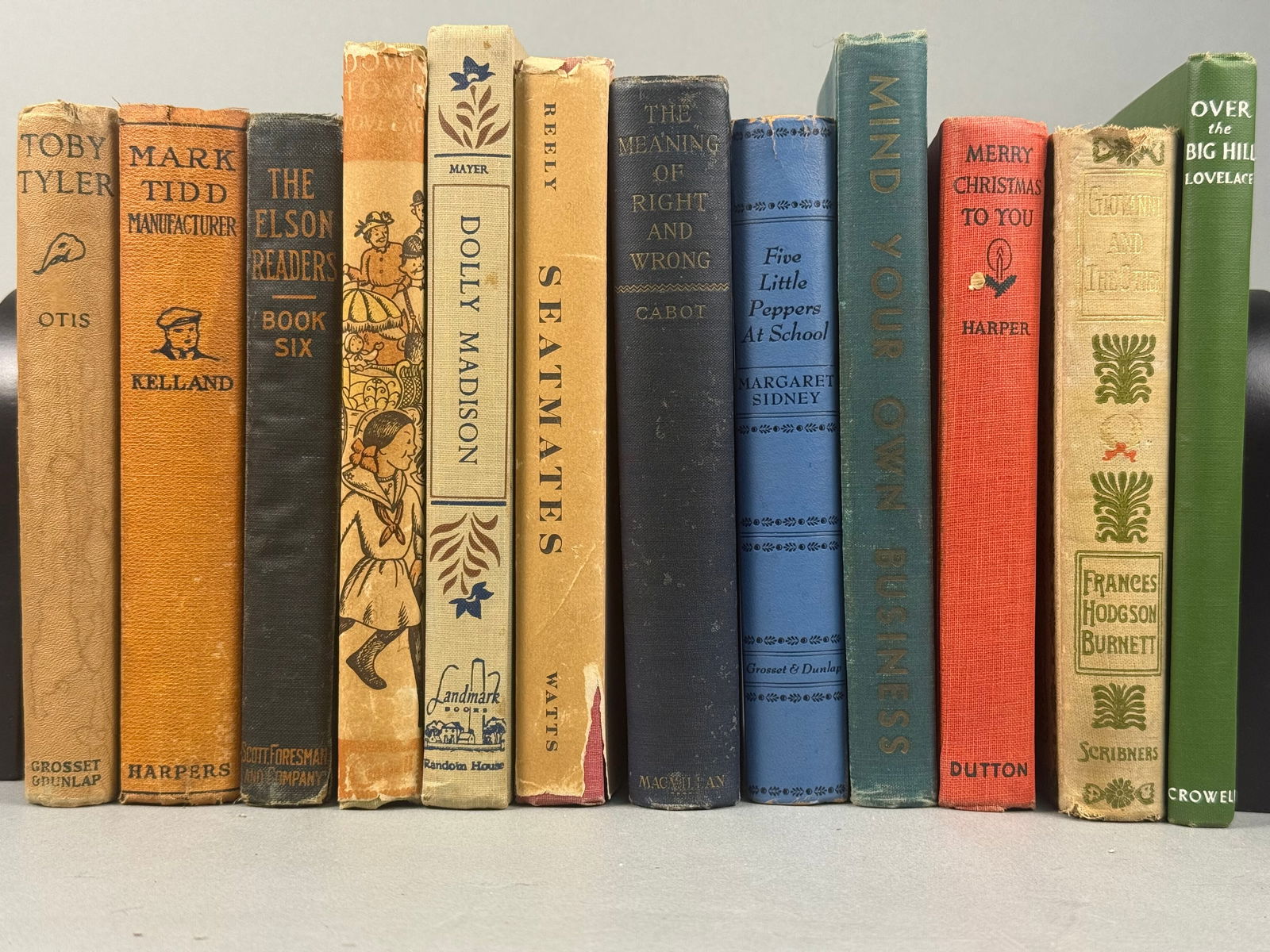 Collection of Vintage Hardcover Books: The bindings feature various cloth and paper over board materials in a spectrum of colors including tan, orange, dark blue, cream, light blue, teal, red, and dark green. Visible titles include "T