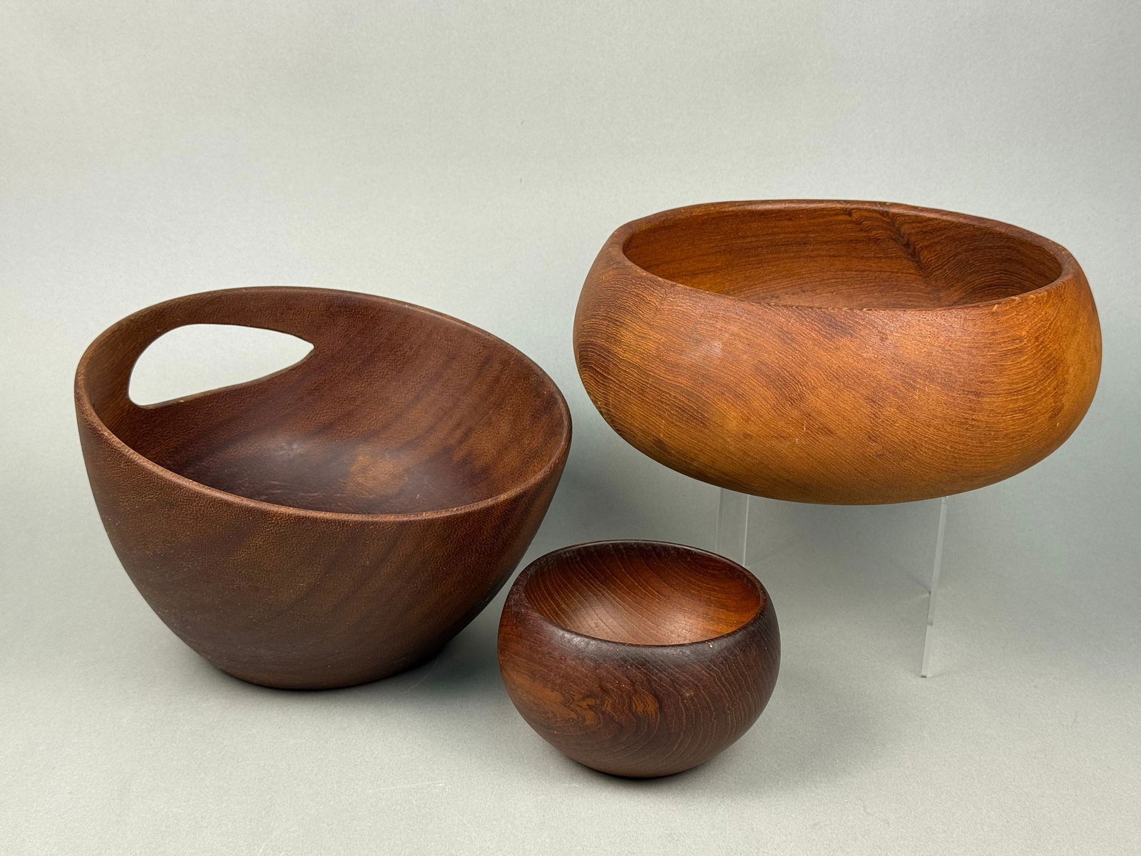 Three Mid-Century Modern Teak Wood Bowls: A set of three Mid-Century Modern teak wood bowls. The collection includes one large, darker brown bowl featuring an integrated handle cut-out, one large, lighter reddish-brown circular bowl, and one
