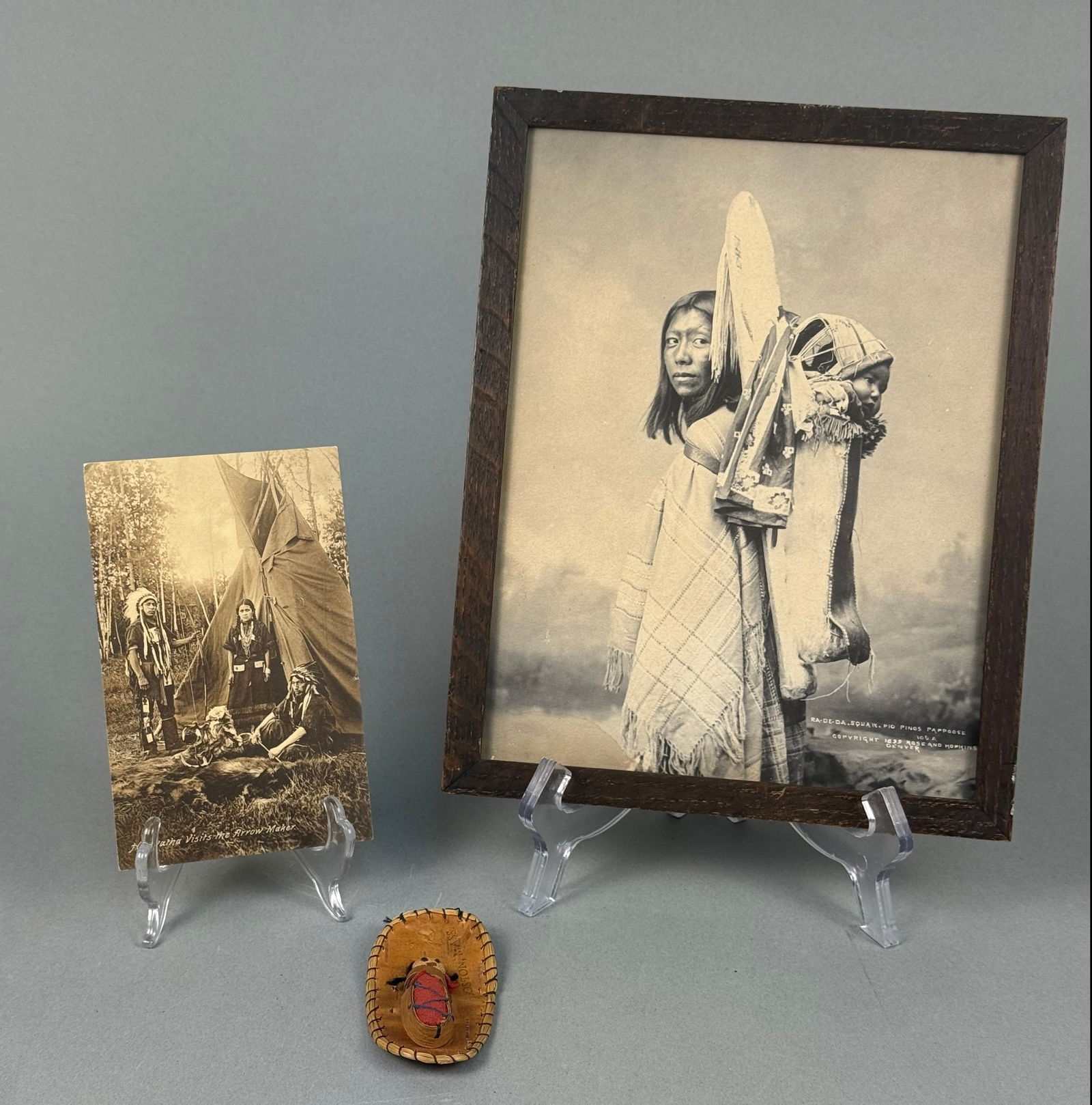 A Group of Three Items: A Rose and Hopkins Photograph of Ra-de-da, an Indigenous Postcard, and a: This lot comprises three items: a framed black and white photograph, a sepia-toned postcard, and a miniature birch bark cradleboard with a small cloth doll. The photograph, depicting an Indigenous wom