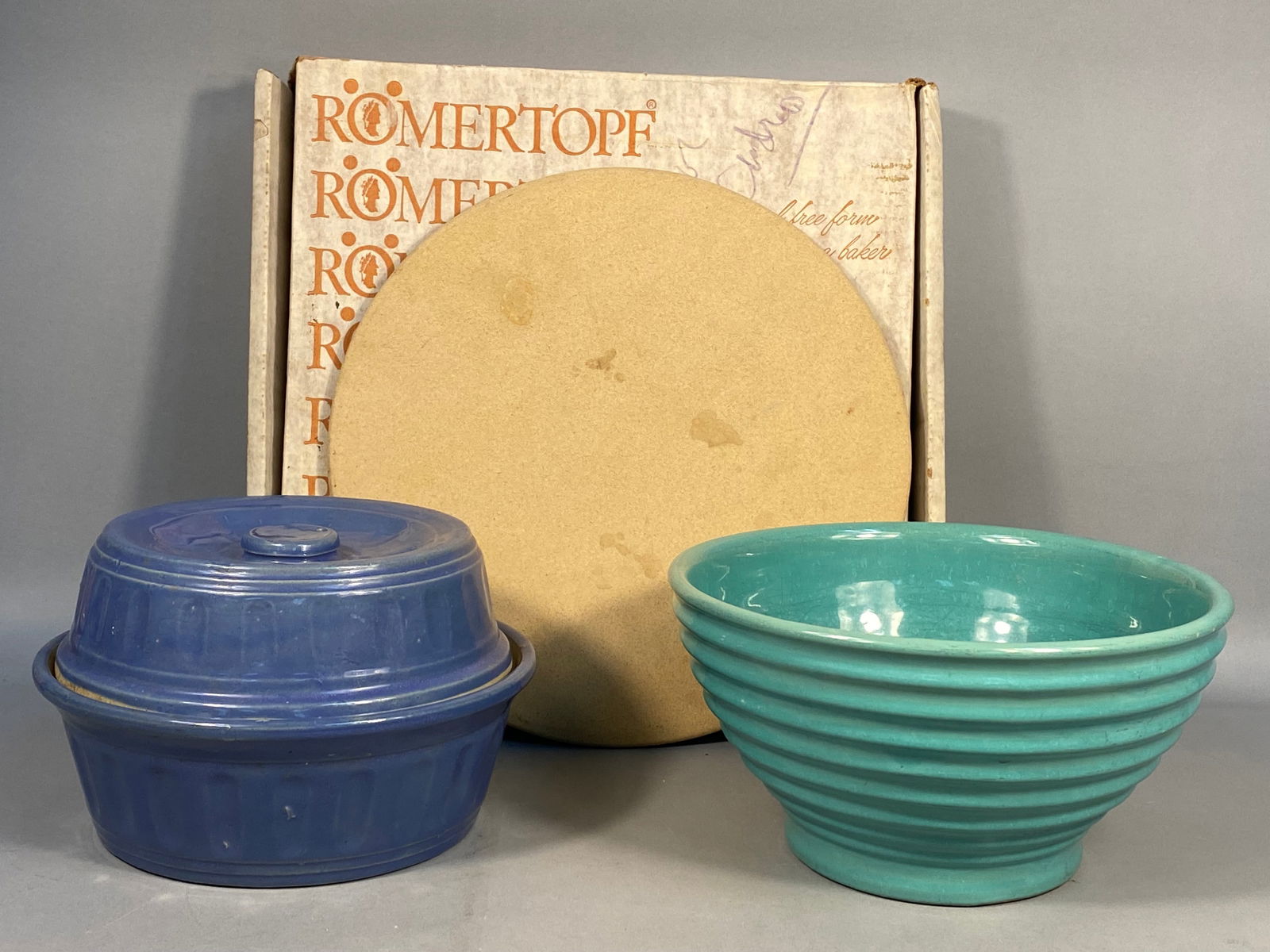 Monmouth Western Stoneware Periwinkle Blue Casserole, a Turquoise Ceramic Bowl, and a R?mertopf: This lot presents a collection of vintage-style kitchenware comprising two glazed ceramic items and an unglazed ceramic baking stone, along with original packaging. The first glazed item is a round, l