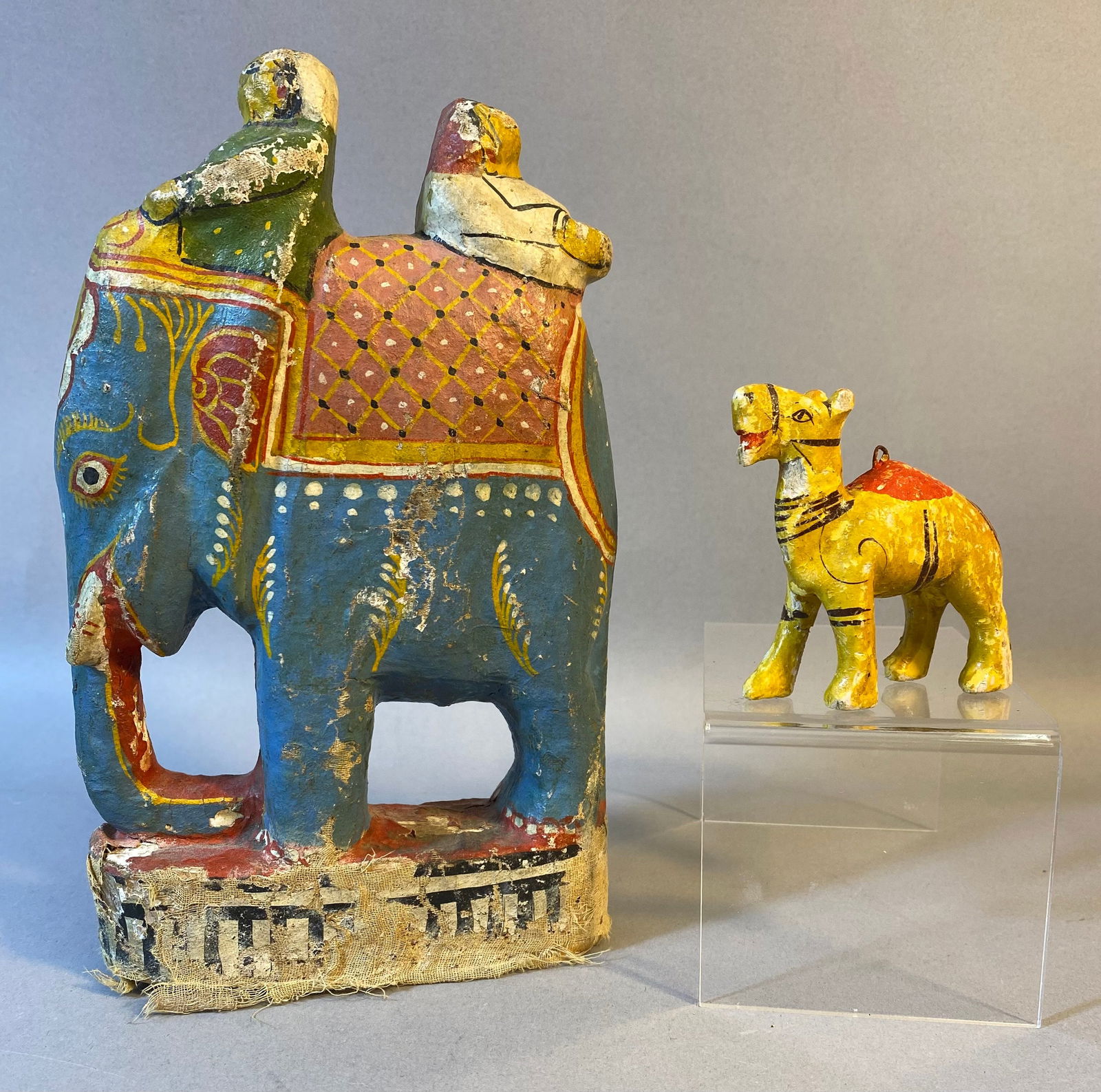 Two Polychrome Painted Folk Art Wood Figurines, an Elephant with Riders and a Camel: The lot comprises two polychrome painted folk art-style figurines. The larger figurine depicts an elephant carrying two stylized riders, while the smaller is a single-humped camel with a small metal l