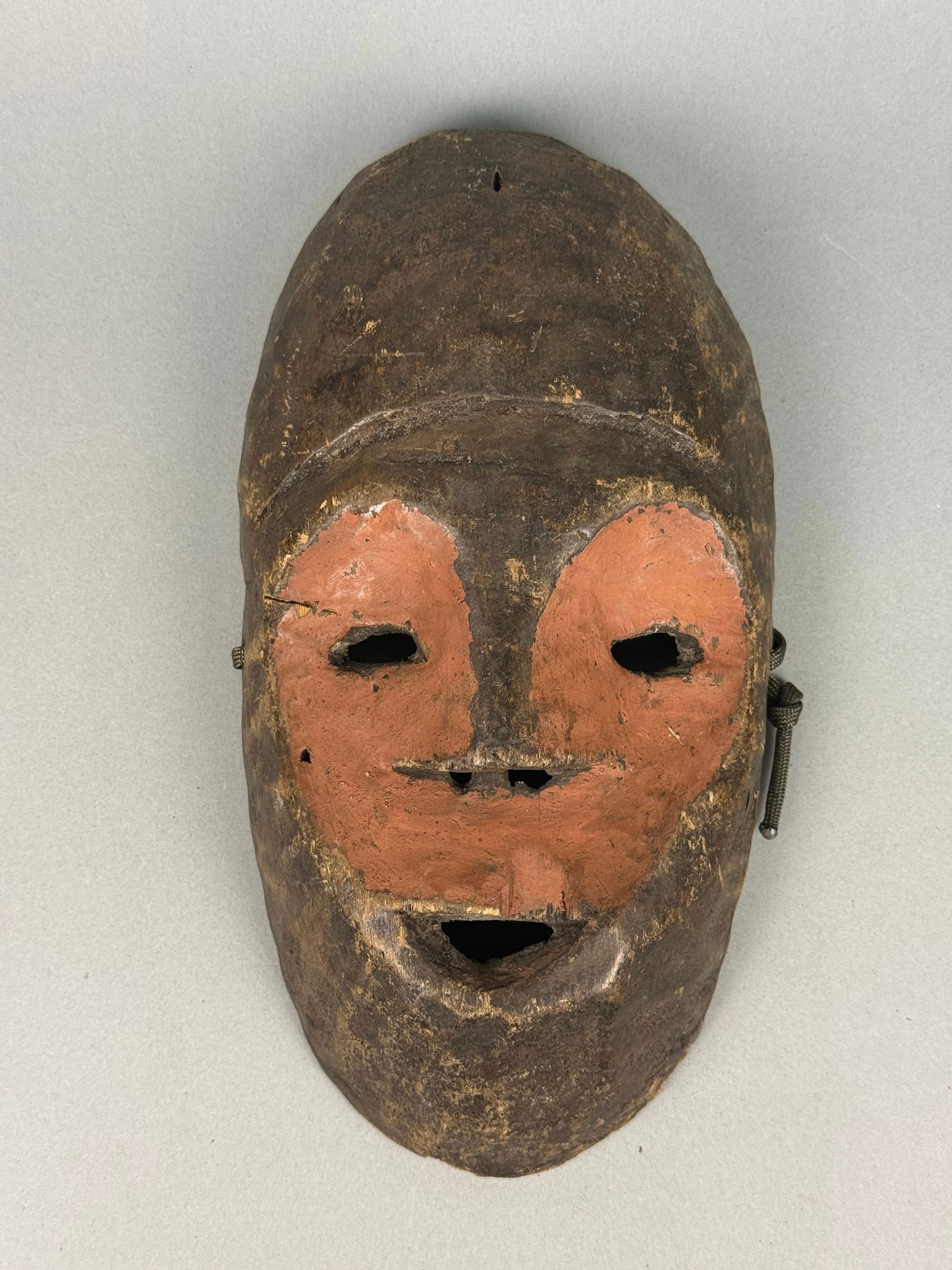 African Tribal Ceremonial Wooden Mask: An African tribal ceremonial wooden mask, depicting a highly stylized human face with open apertures for the eyes and mouth, and a prominent, projecting nose. The mask features a dark, deeply patinate