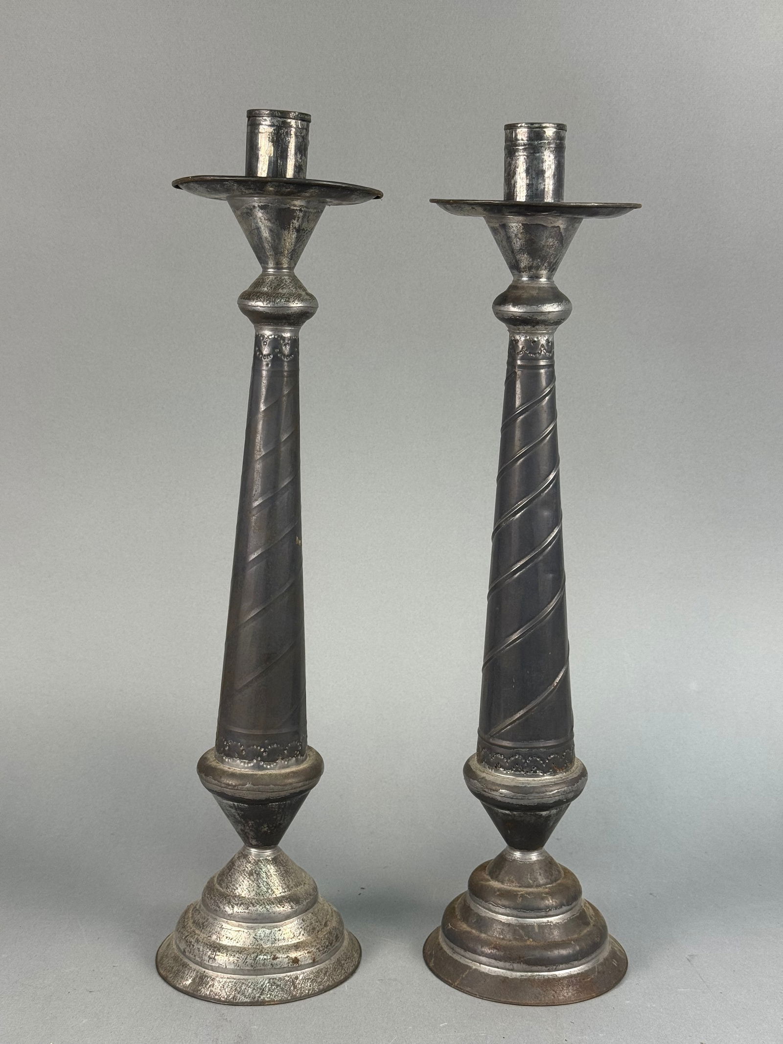 A Pair of Large Mexican Hojalata Tin Candlesticks: A pair of large Mexican Hojalata tin candlesticks. Each candlestick features a cylindrical candle holder above a shallow drip pan, connected to a tall, tapering shaft with a pronounced spiral fluting.