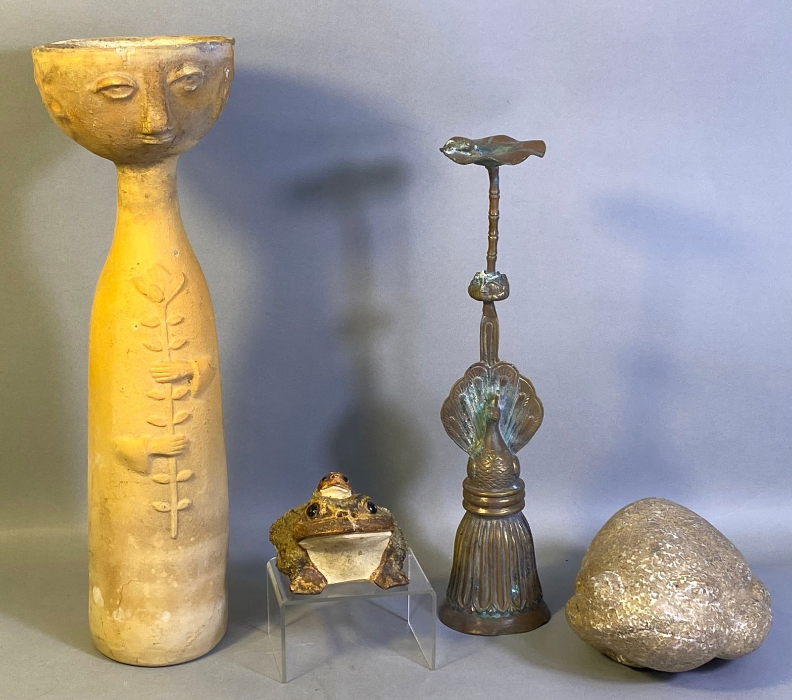 A Group of Four Decorative Objects, including an Earthenware Vessel and a Brass Candlestick: A group of four distinct decorative objects. Comprising: a tall unglazed earthenware or terracotta vessel with a narrow body and a stylized human face forming the rim, adorned with relief hands holdin