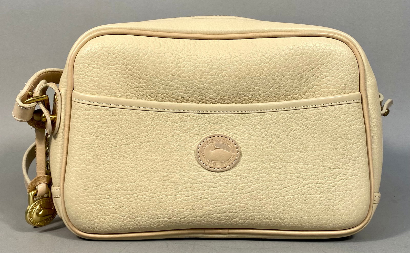 Vintage Dooney & Bourke All-Weather Leather Cream Pebbled Leather Crossbody Bag: A Vintage Dooney & Bourke All-Weather Leather crossbody bag. This rectangular cream-colored bag is constructed from pebbled leather with smooth light beige leather trim and features an adjustable stra