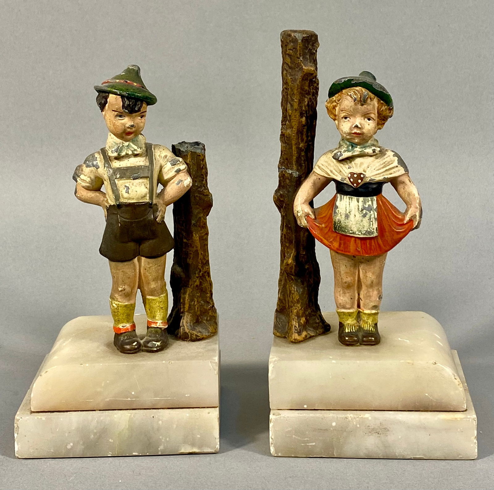 A Pair of Early 20th Century Austrian Cast Metal Alpine Children Bookends: A pair of early 20th century figural bookends, each featuring a hand-painted cast metal figure, likely spelter, depicting a child in traditional Alpine attire. The boy is dressed in lederhosen and the